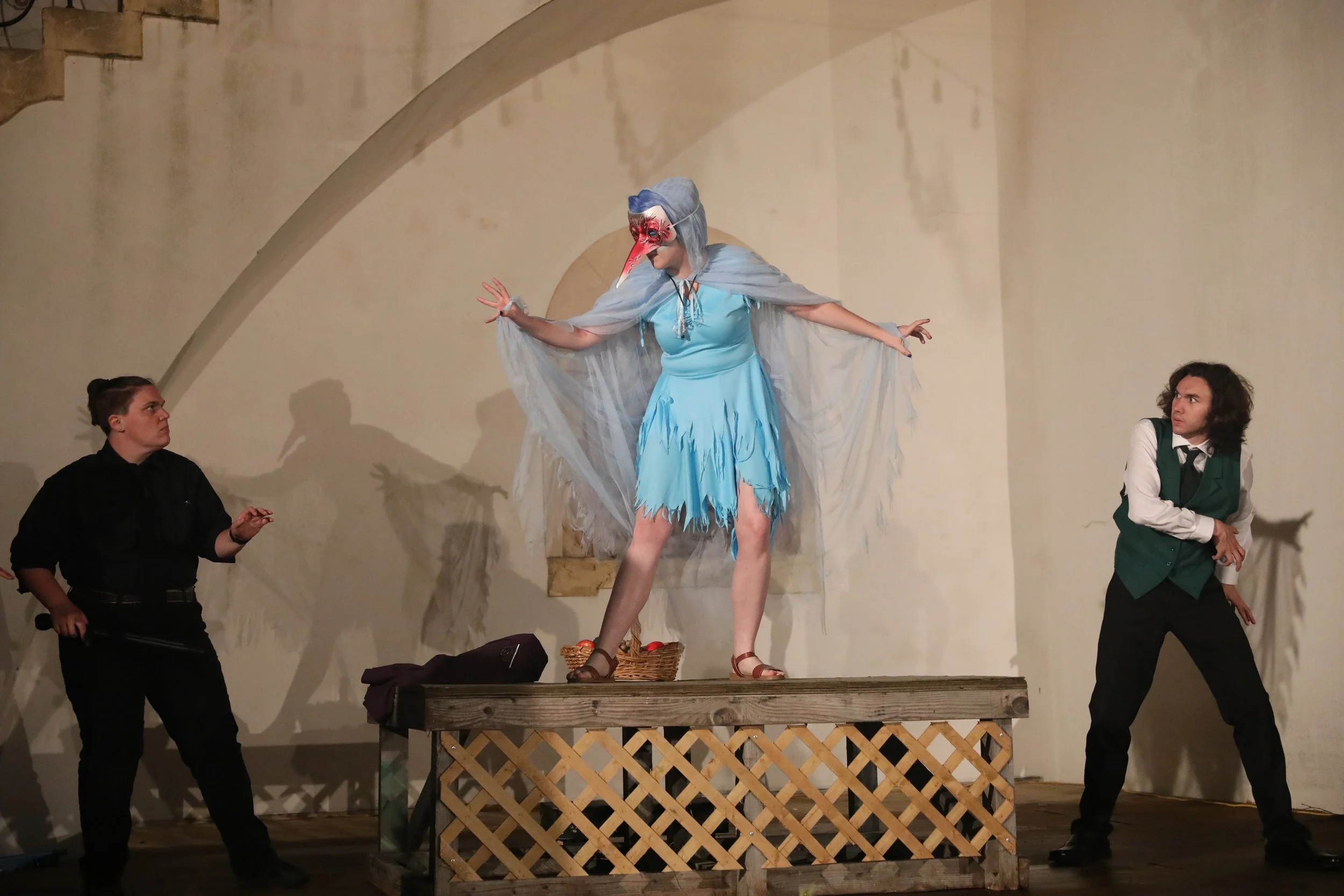A theatrical performance featuring a woman wearing a blue dress, a blue hair wig, a red carnival mask with a long nose, and tattered blue fabric draped over her. She is standing on a wooden platform with arms outstretched.