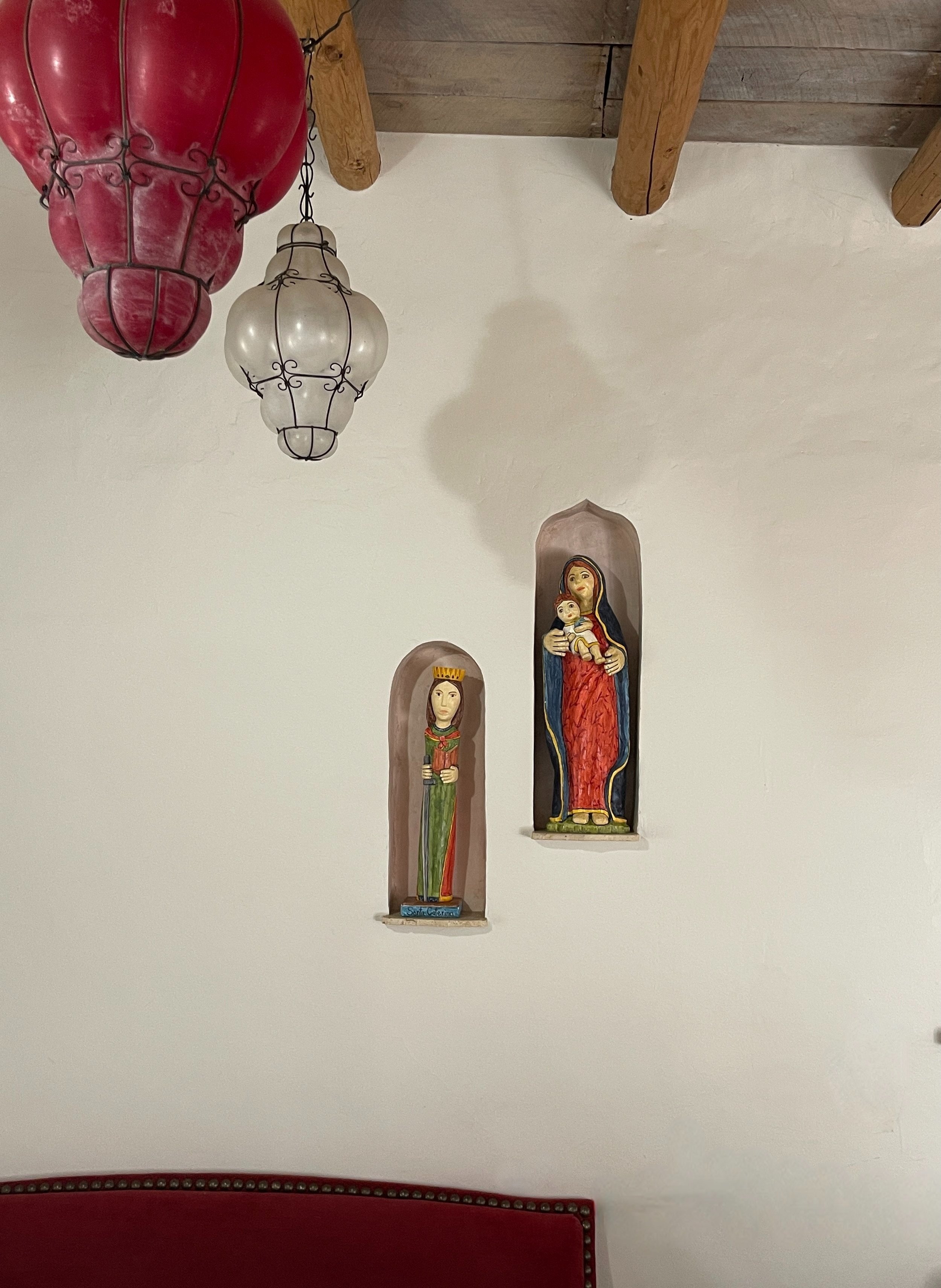 Colorful statues of religious figures on a white wall next to hanging red and white lamps. Traditional Spanish folk art. These Bultos are hand carved and painted, one of a kind pieces of art, by Barbara Hernandez