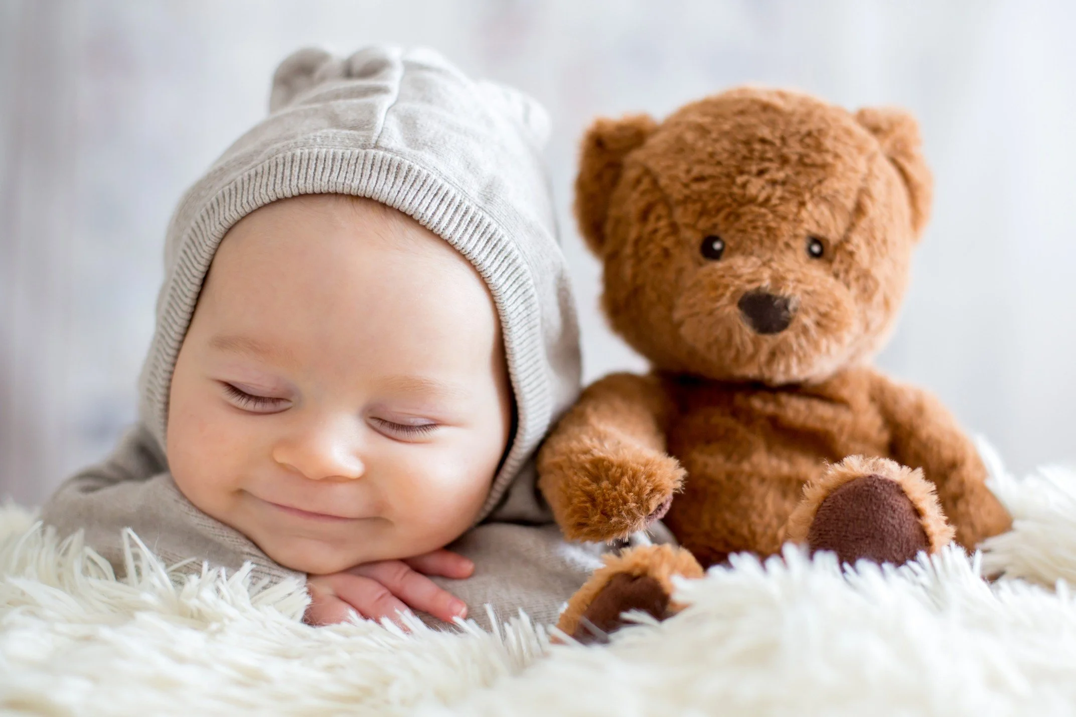 A sleeping baby wearing a gray beanie, resting on a fluffy white rug, with a brown teddy bear sitting beside them.