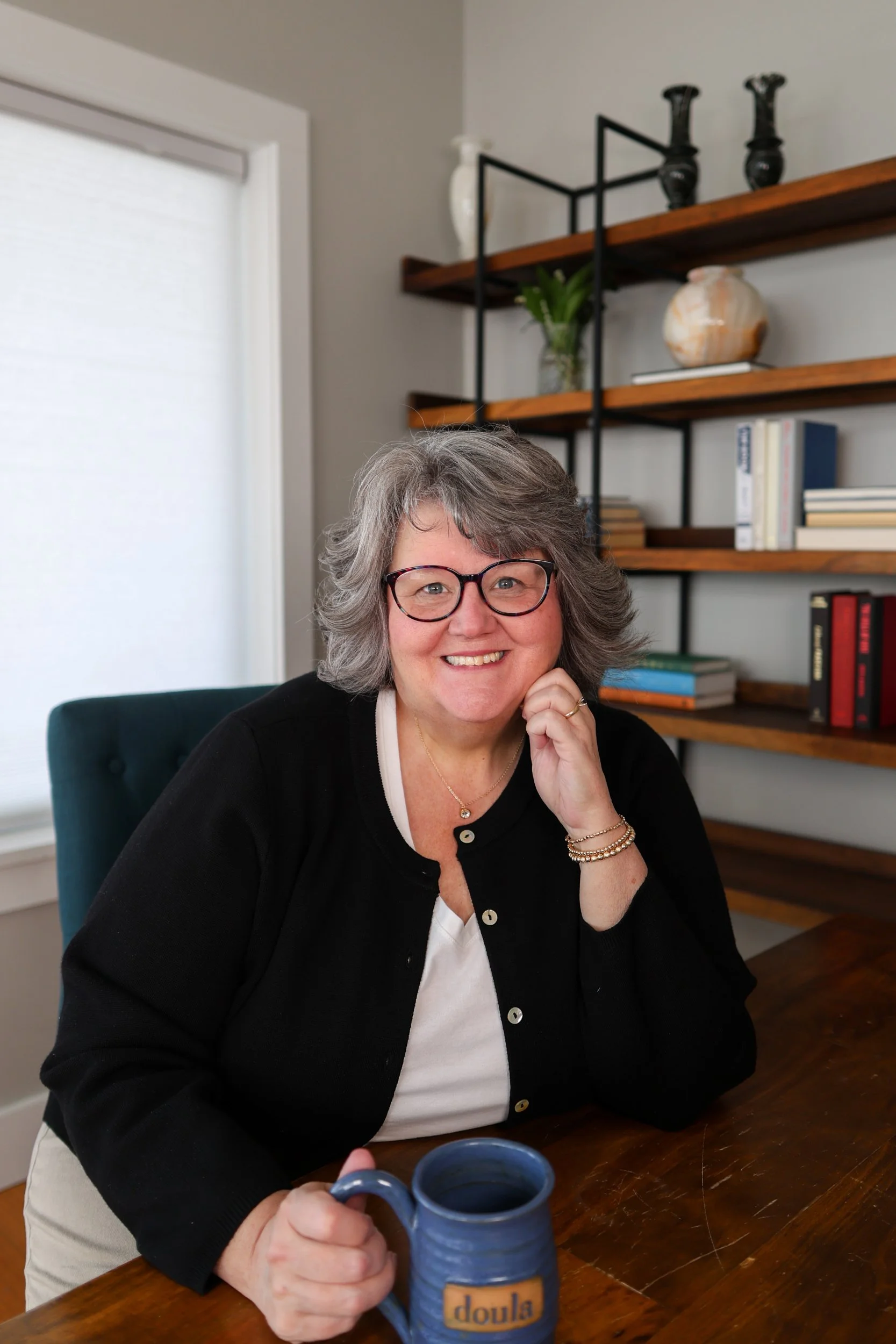 A woman with gray hair, glasses, and a black cardigan smiling at the camera while sitting at a wooden table with a blue mug in her hand. Behind her, there are wooden shelves with decorative items and books, and a window with white blinds.
