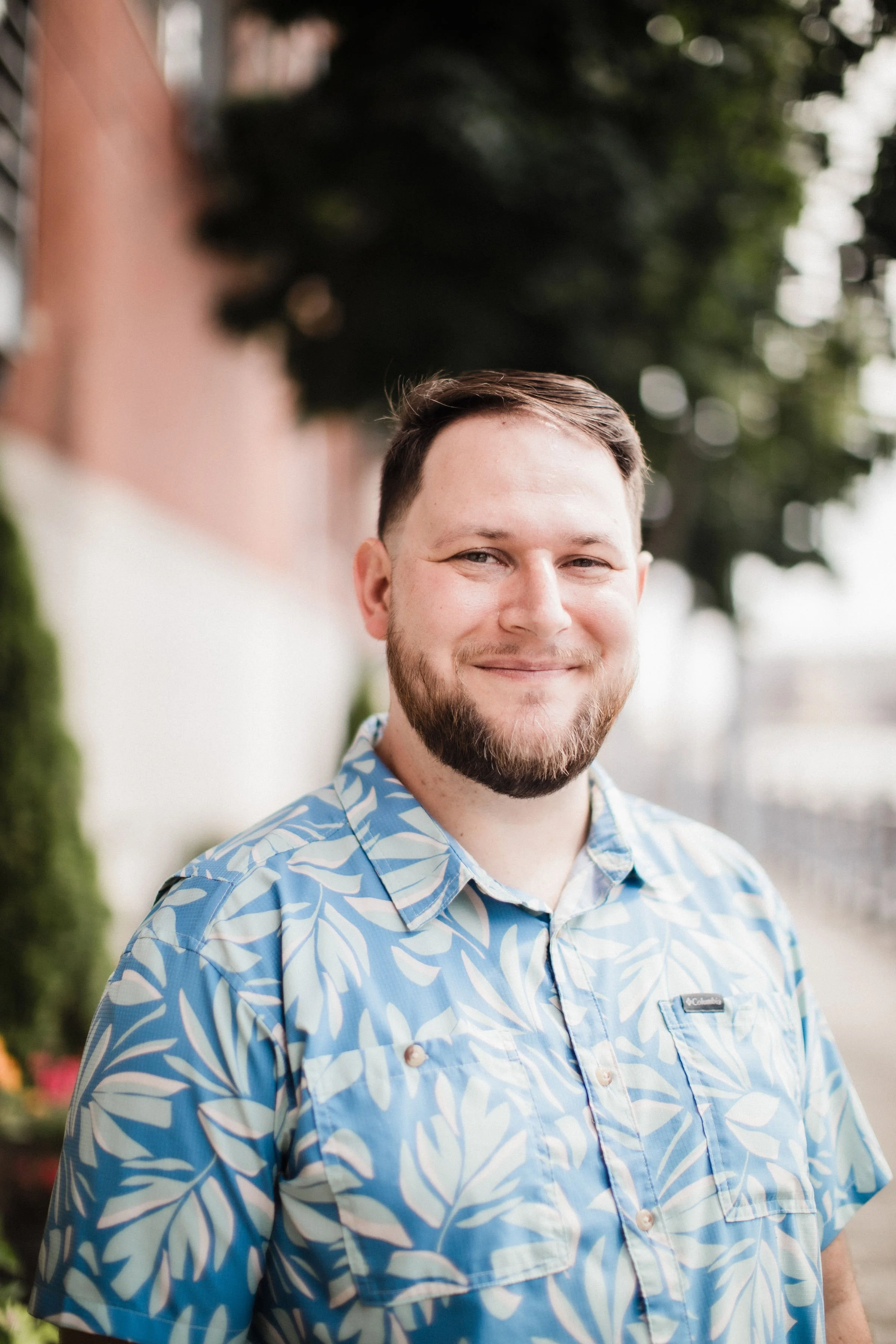 A man wearing a checkered shirt standing outdoors with trees in the background.
