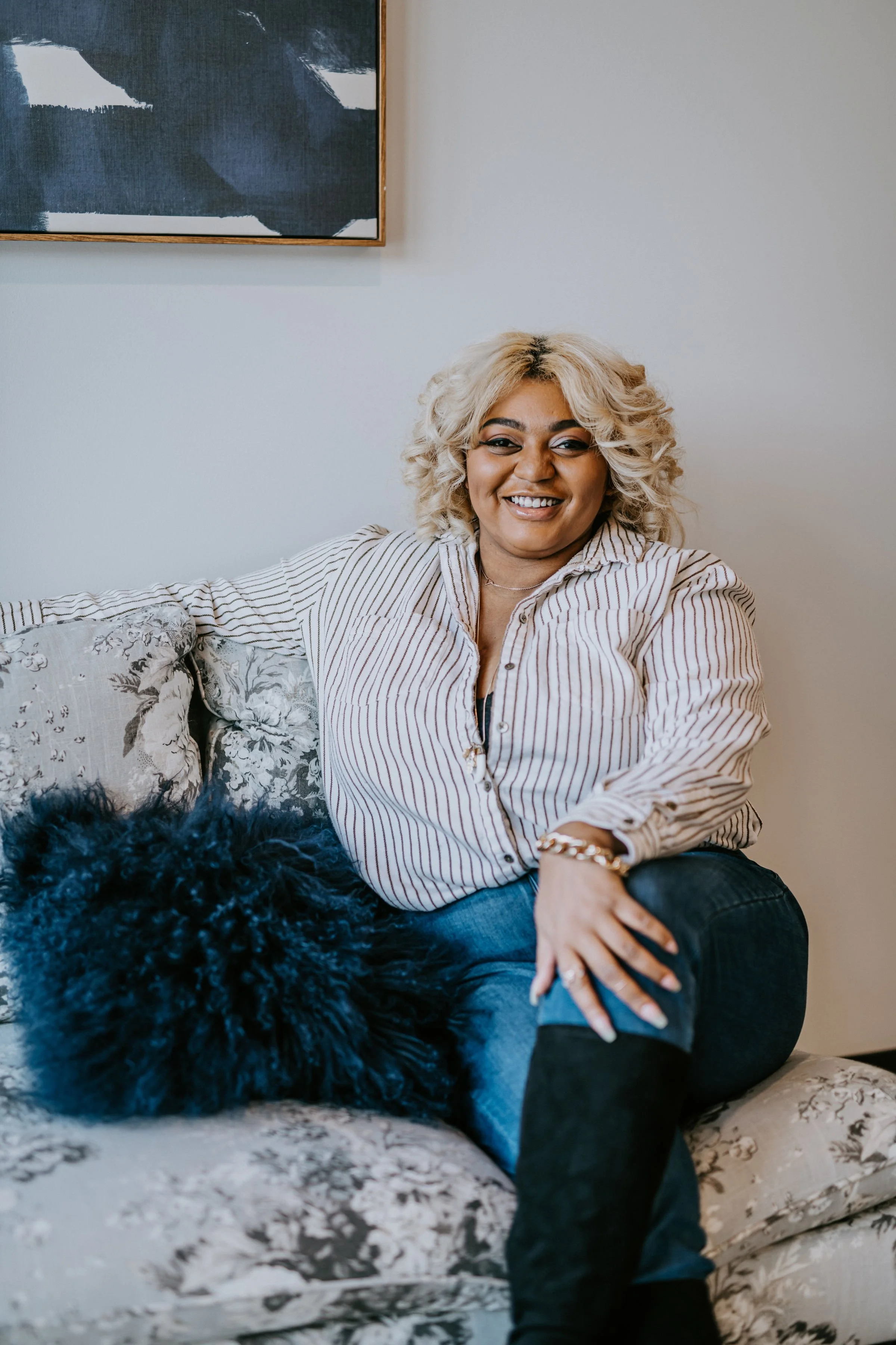 A person with curly blonde hair and a striped shirt smiling while sitting on a floral-patterned couch with a blue fluffy pillow. A piece of abstract art is partially visible on the wall behind them.