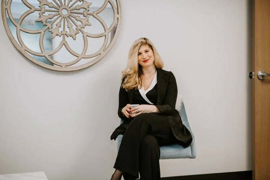 A woman with long blonde hair wearing a black suit sits on a chair in a modern office setting, smiling. A round decorative mirror is mounted on the wall beside her, and a wooden door is visible on the right.