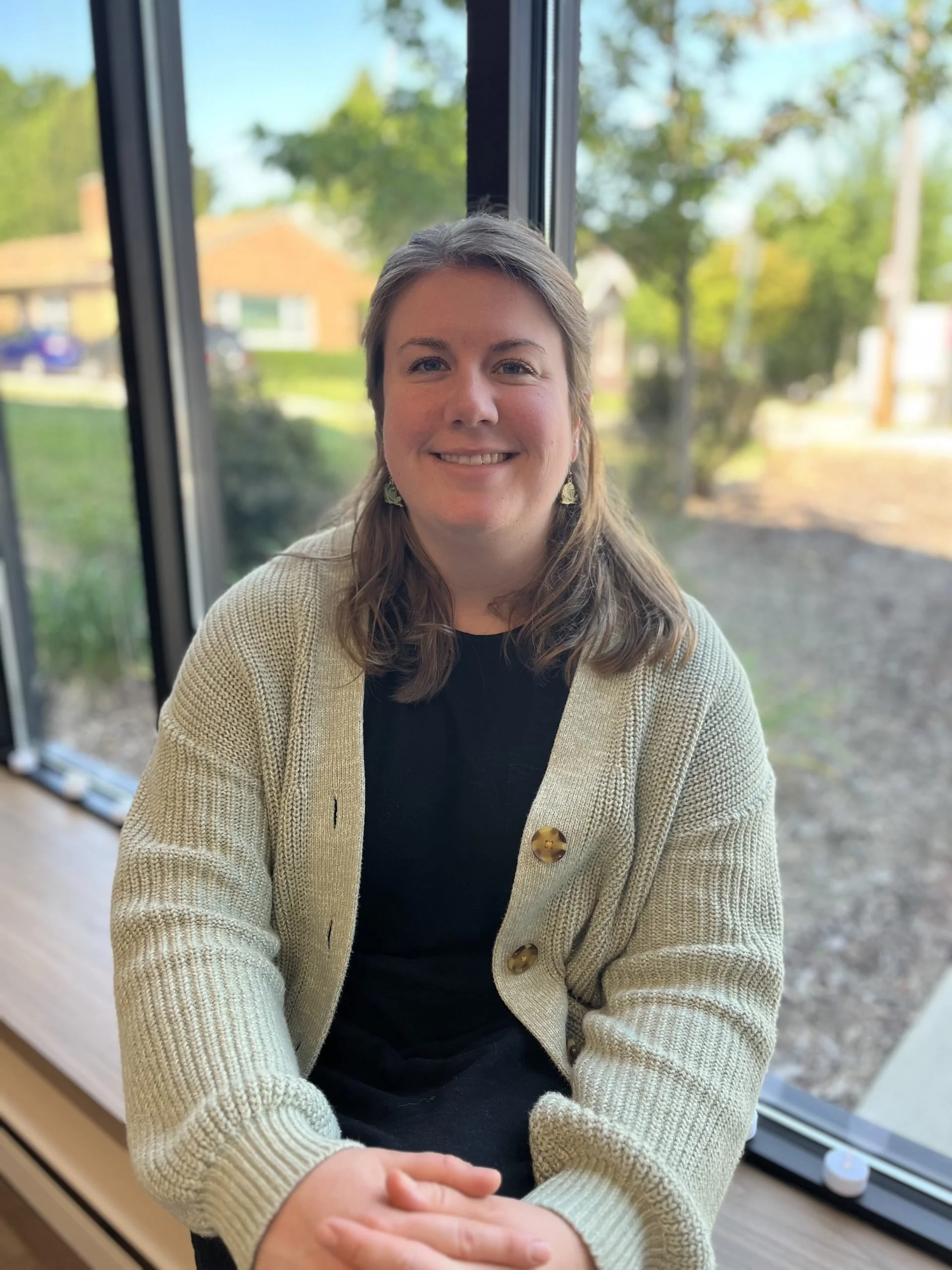 Smiling person sitting by a window with trees and houses visible outside.