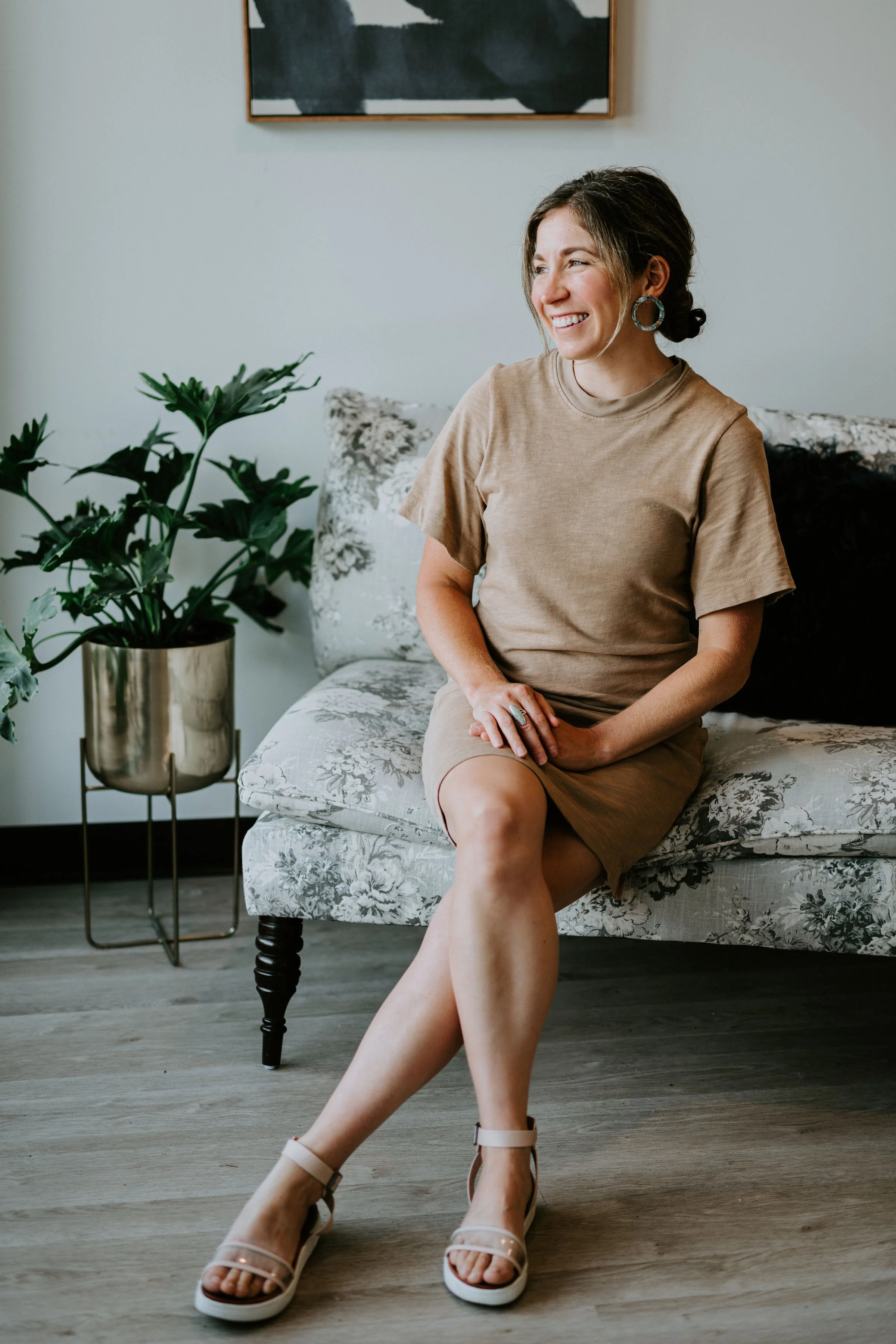 Woman sitting on a patterned sofa with a potted plant nearby, wearing a beige dress and sandals, in a modern indoor setting.