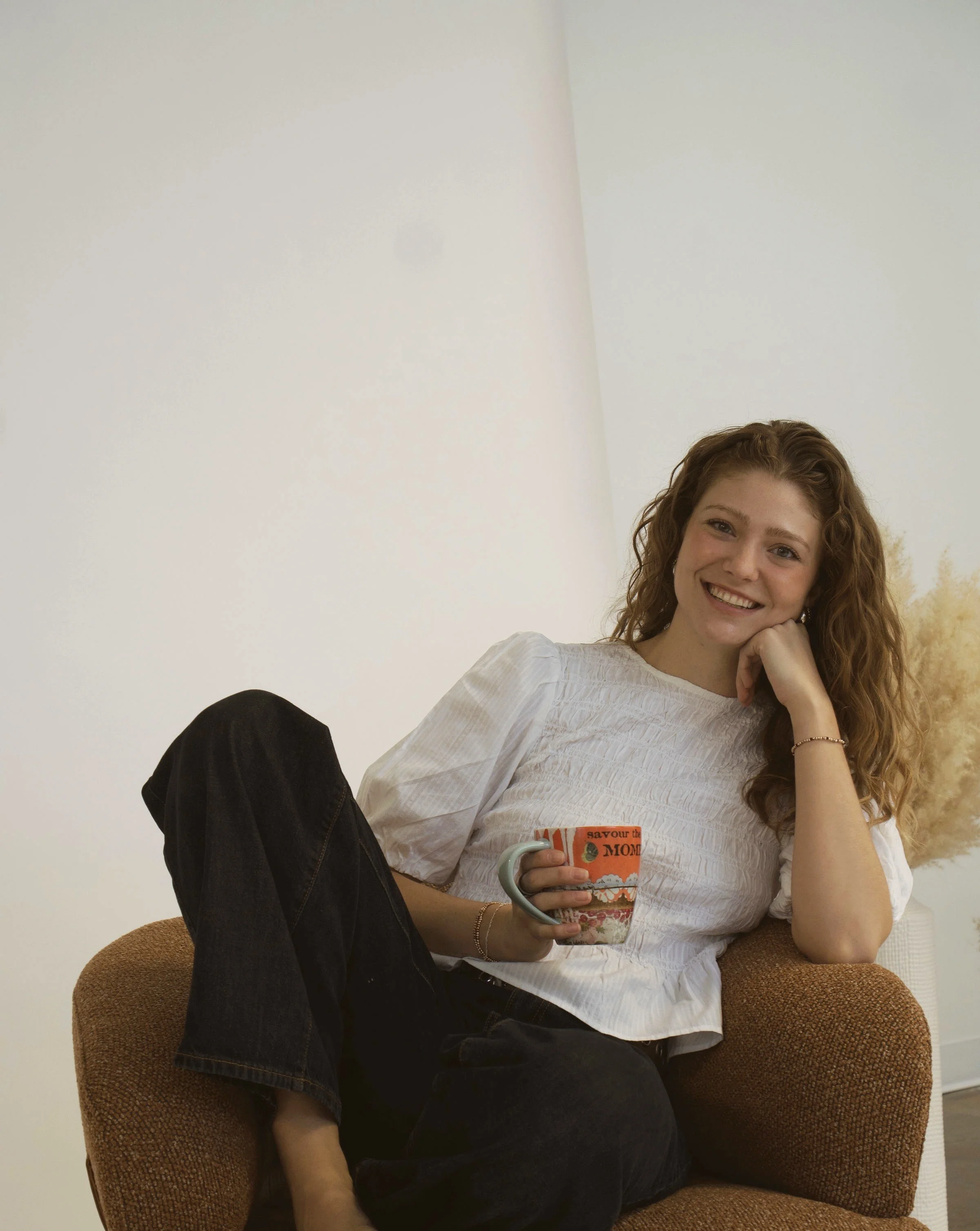 A young woman with curly red hair, sitting comfortably on an armchair, smiling, holding a colorful mug, wearing a white blouse and black pants.
