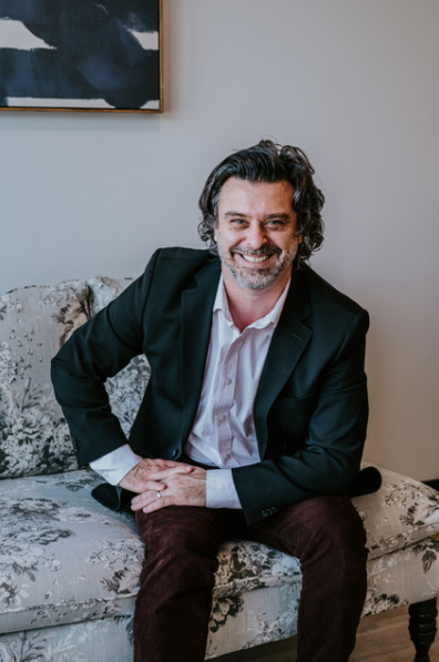 Man sitting on a floral-patterned sofa, wearing a black blazer, white shirt, and dark pants, smiling.
