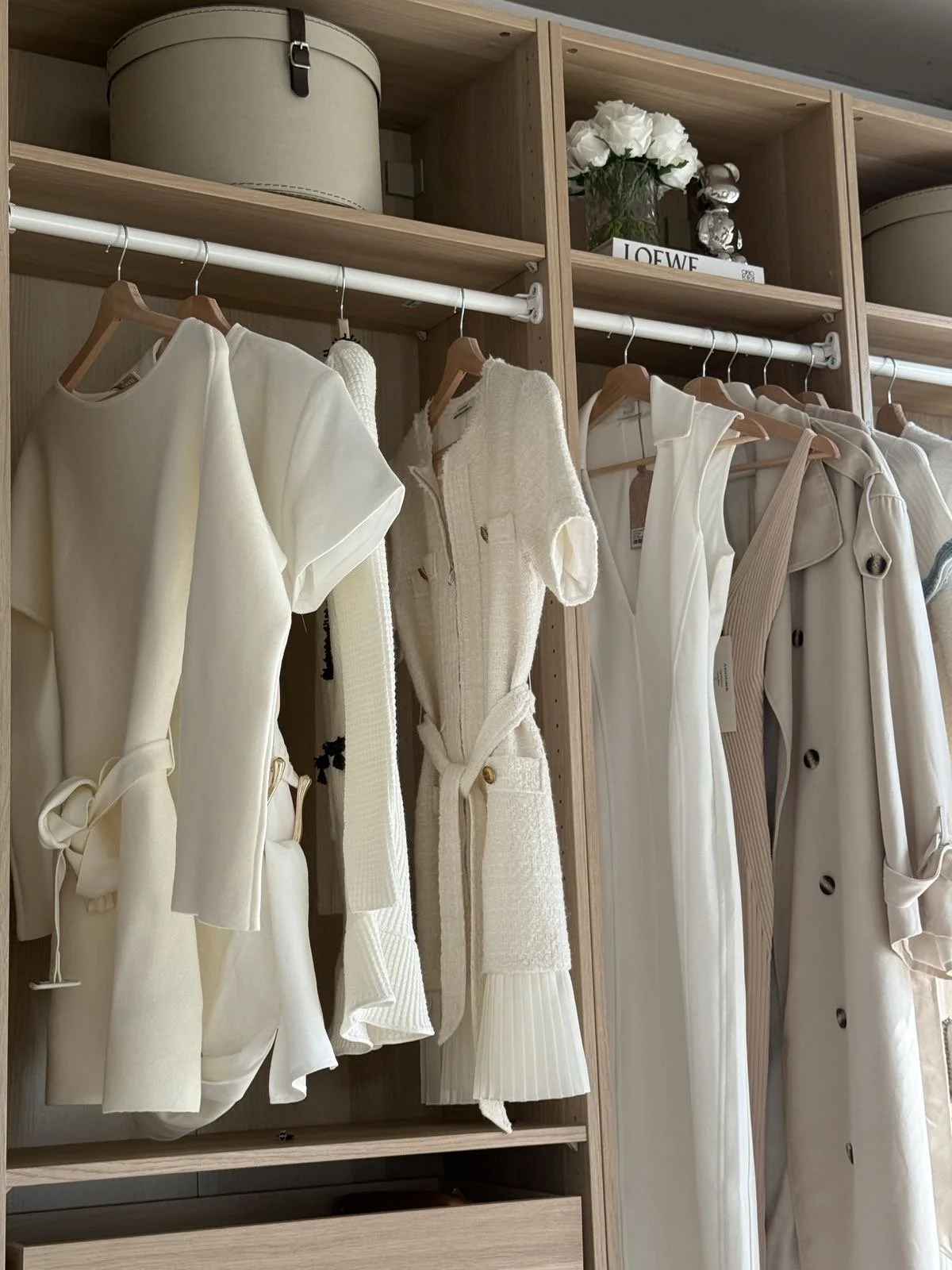 Closet with white and beige clothing hanging on wooden hangers, including dresses, vests, and coats, with wooden shelves above holding decorative boxes, a vase of white roses, and a Loewe box.