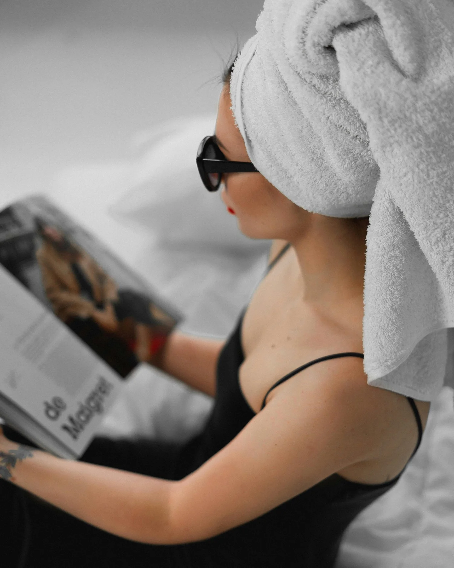 Woman with towel on her head and sunglasses reading a magazine in bed.