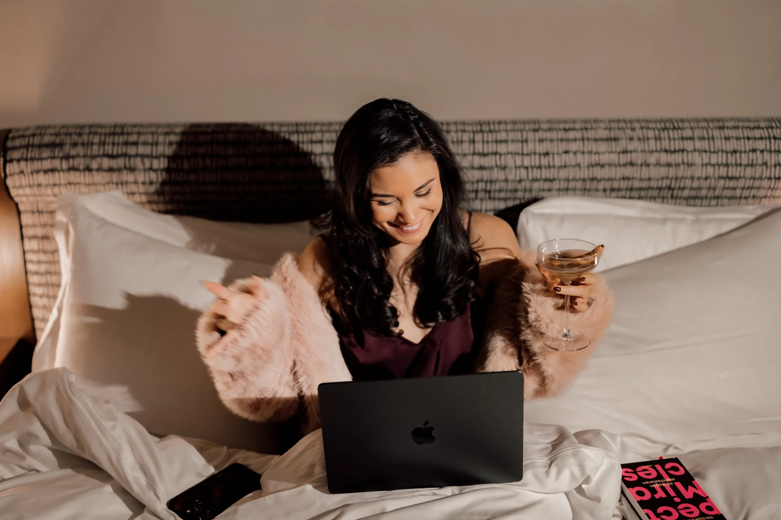 Vrouw zit op bed met een laptop, drinkt wijn, glimlacht en heeft een boek en smartphone naast zich.