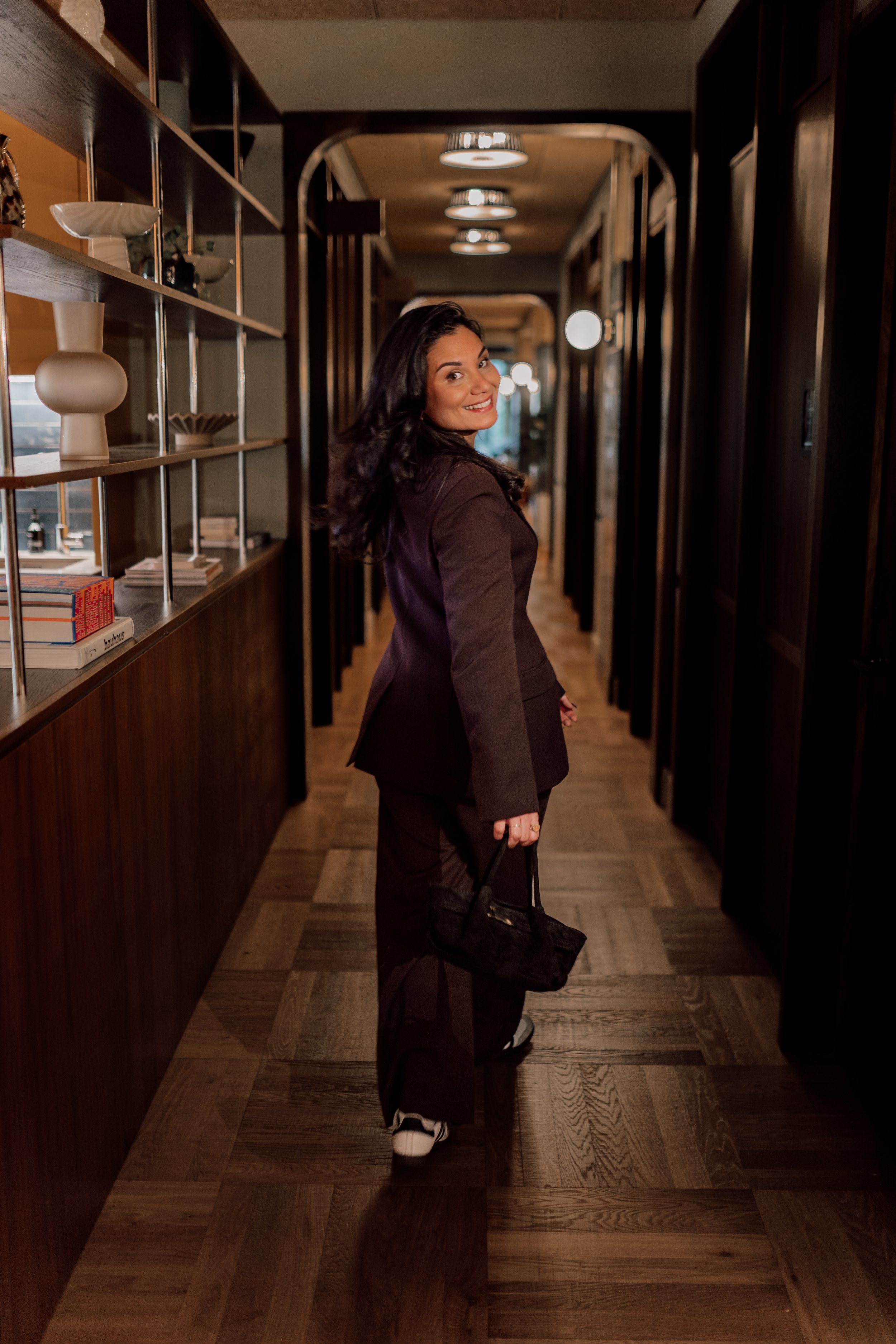 Vrouw in een donkerbruin pak en witte sneakers in een interieur met houten vloeren en donkere houten panelen, lachend naar de camera terwijl ze een zwart tasje vasthoudt.