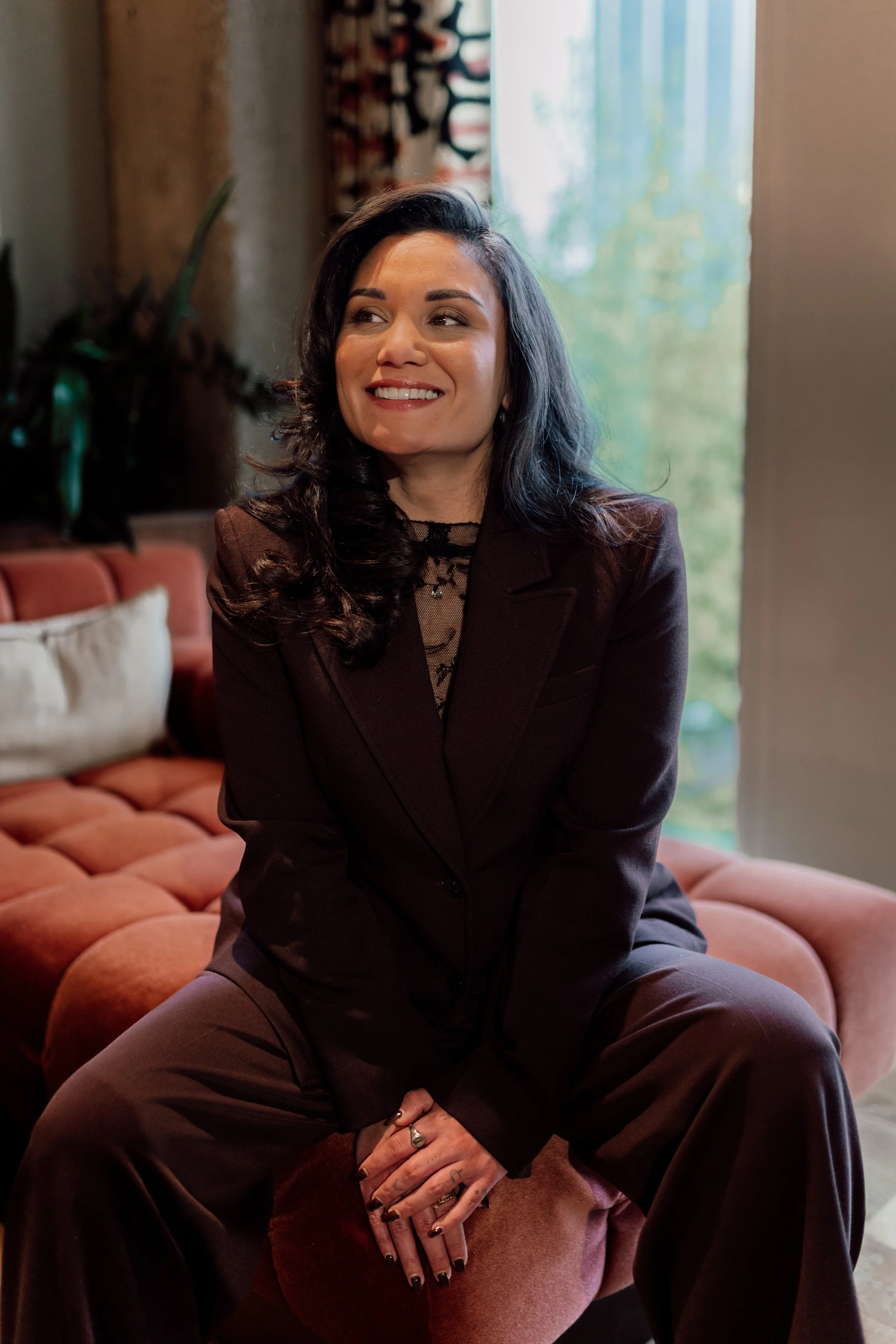 Vrouw met zwarte haar in een bruin pak, zittend op een roze bank in een gezellig interieur, lachend en naar links kijkend.