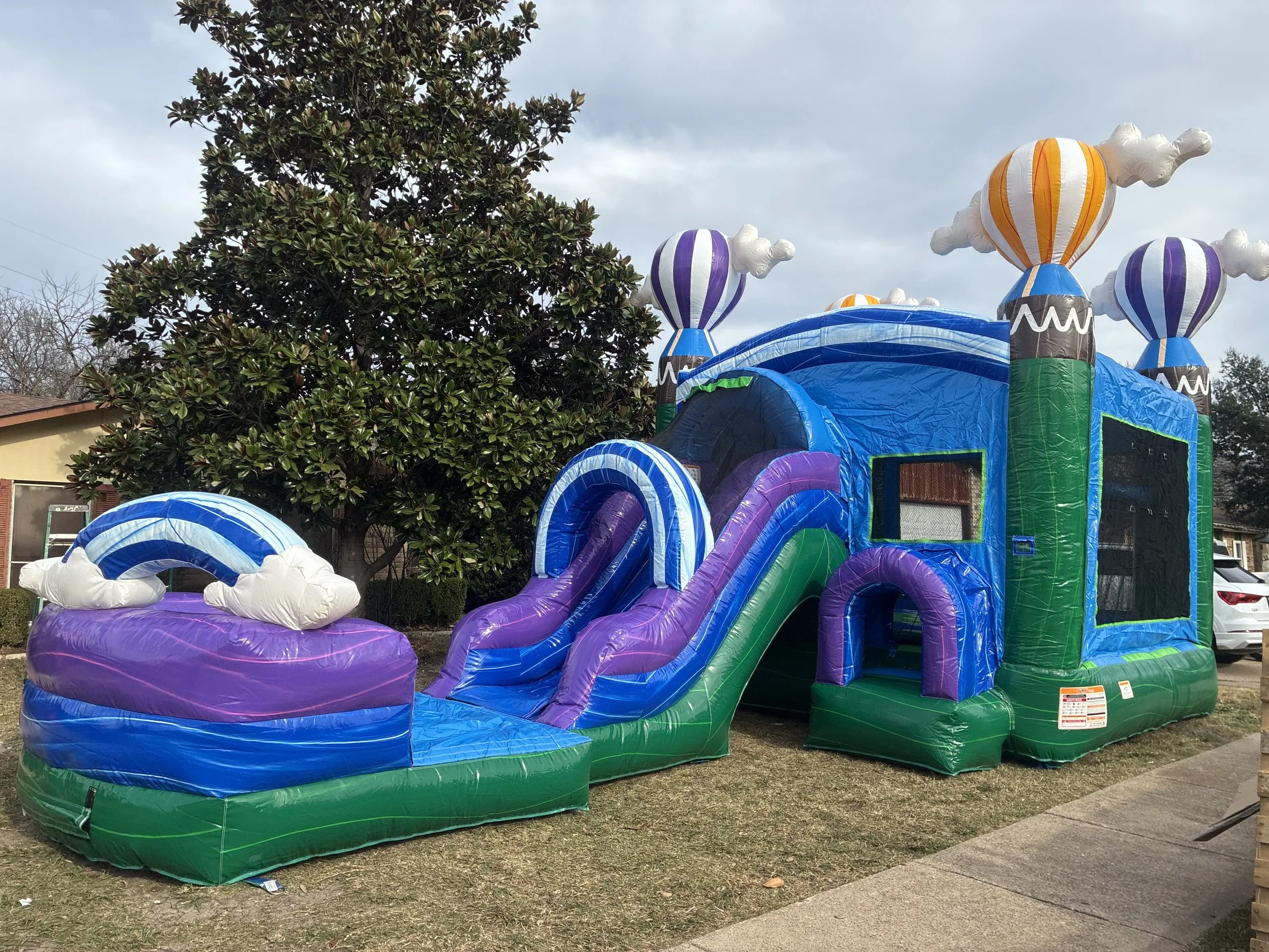 Colorful inflatable bounce house with a slide and hot air balloon decorations on top, set up outdoors on green turf.