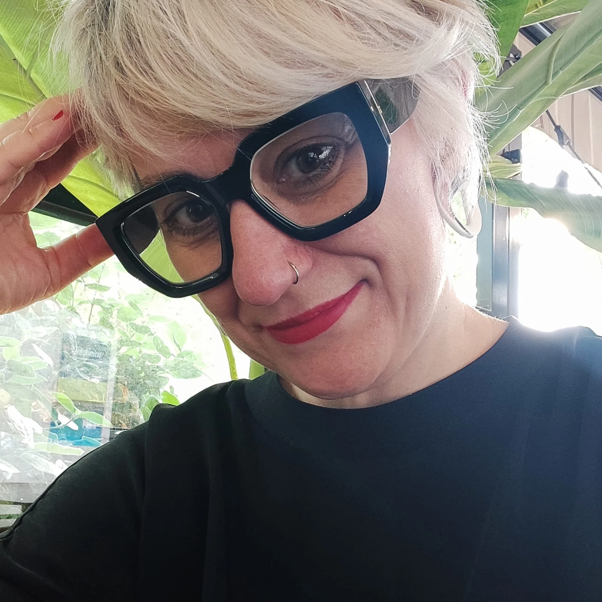 Mujer con gafas negras, rubia y con labios rojos sonriendo cerca de plantas verdes.