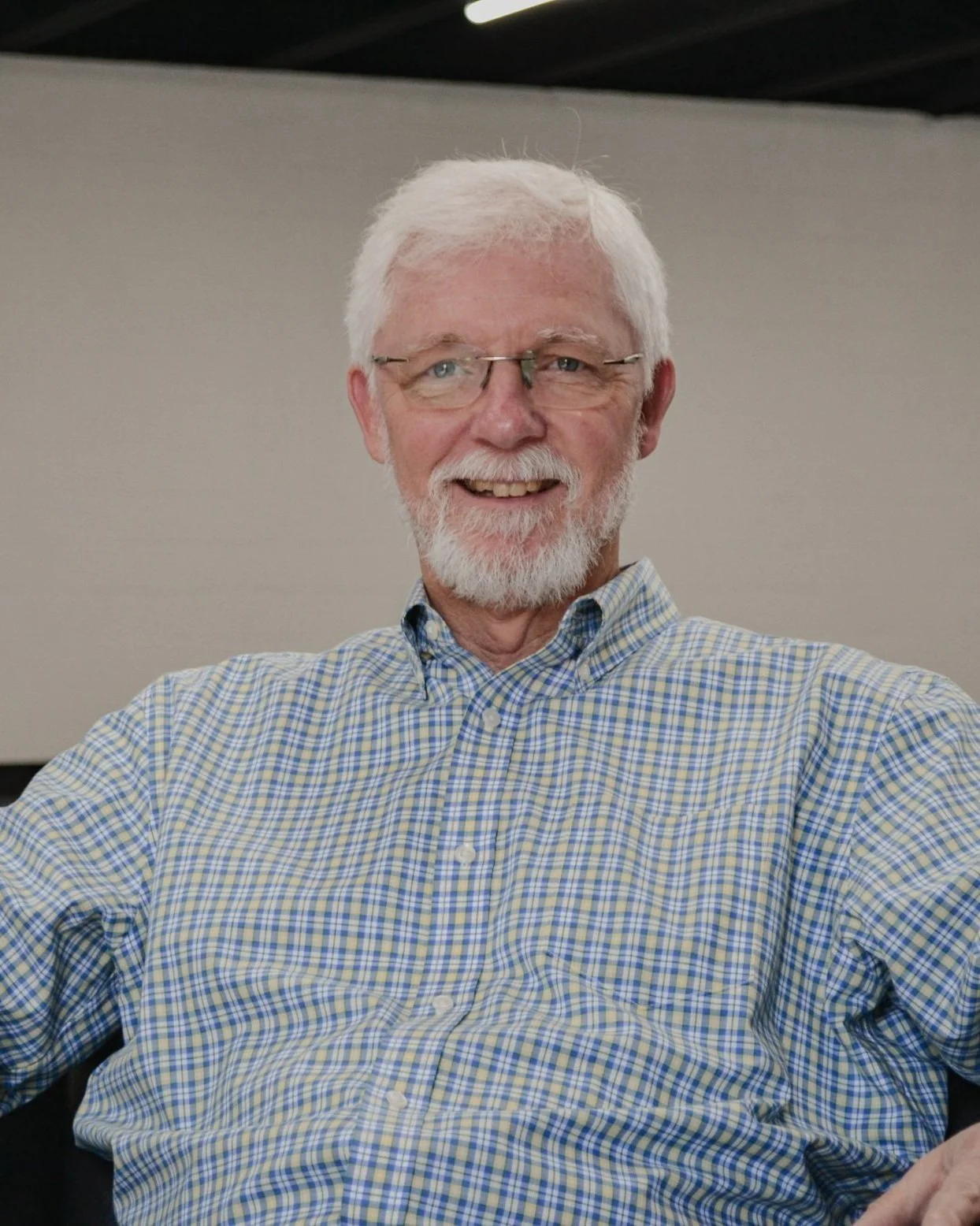 A smiling man with white hair, glasses, and a beard, wearing a plaid shirt, sitting indoors against a plain wall. Casa del Lago's director of adult education.