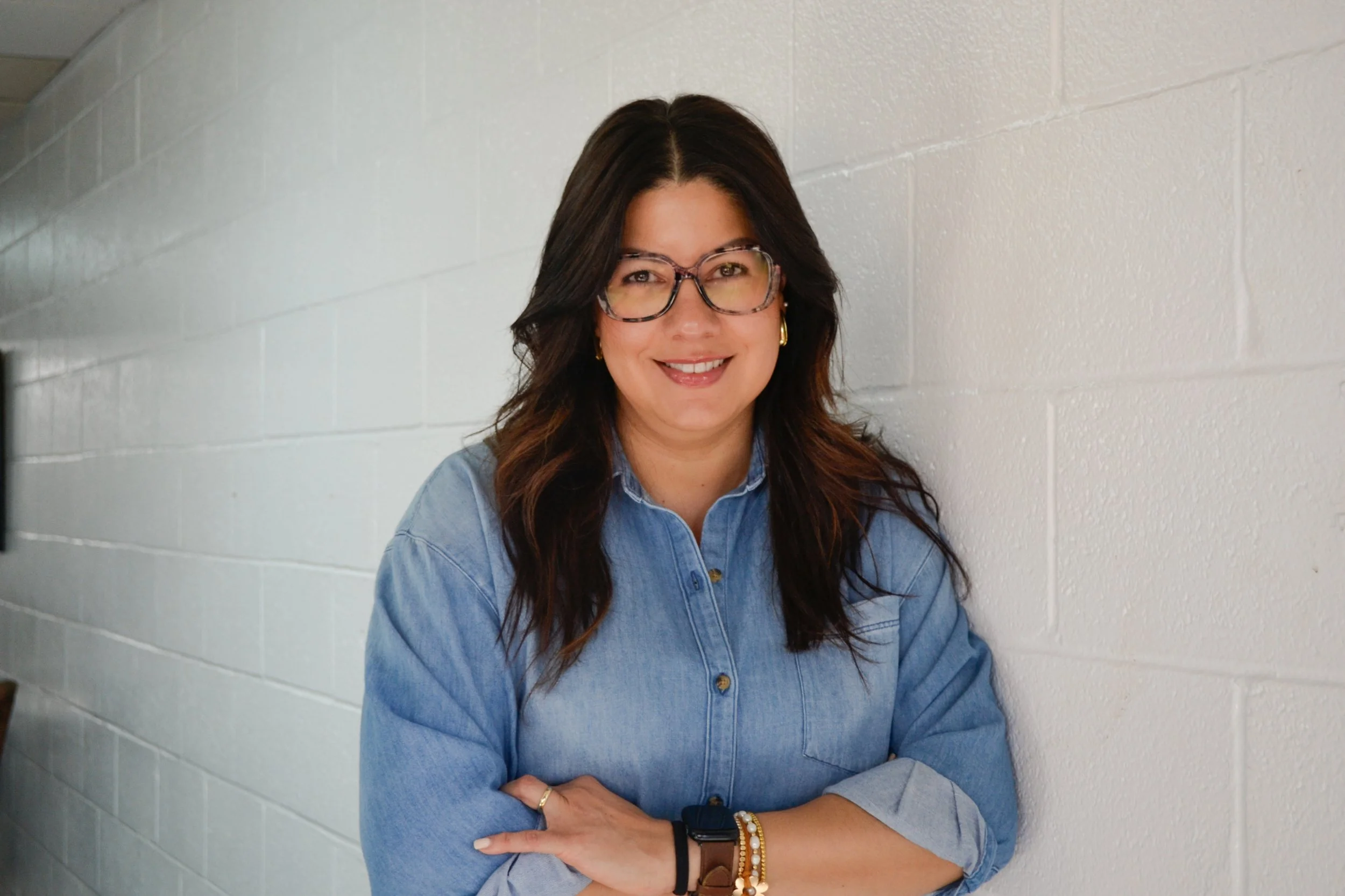 A woman with long dark hair, glasses, and gold earrings posing against a white brick wall, wearing a blue denim shirt with her arms crossed, smiling. Casa del lago's director of operations