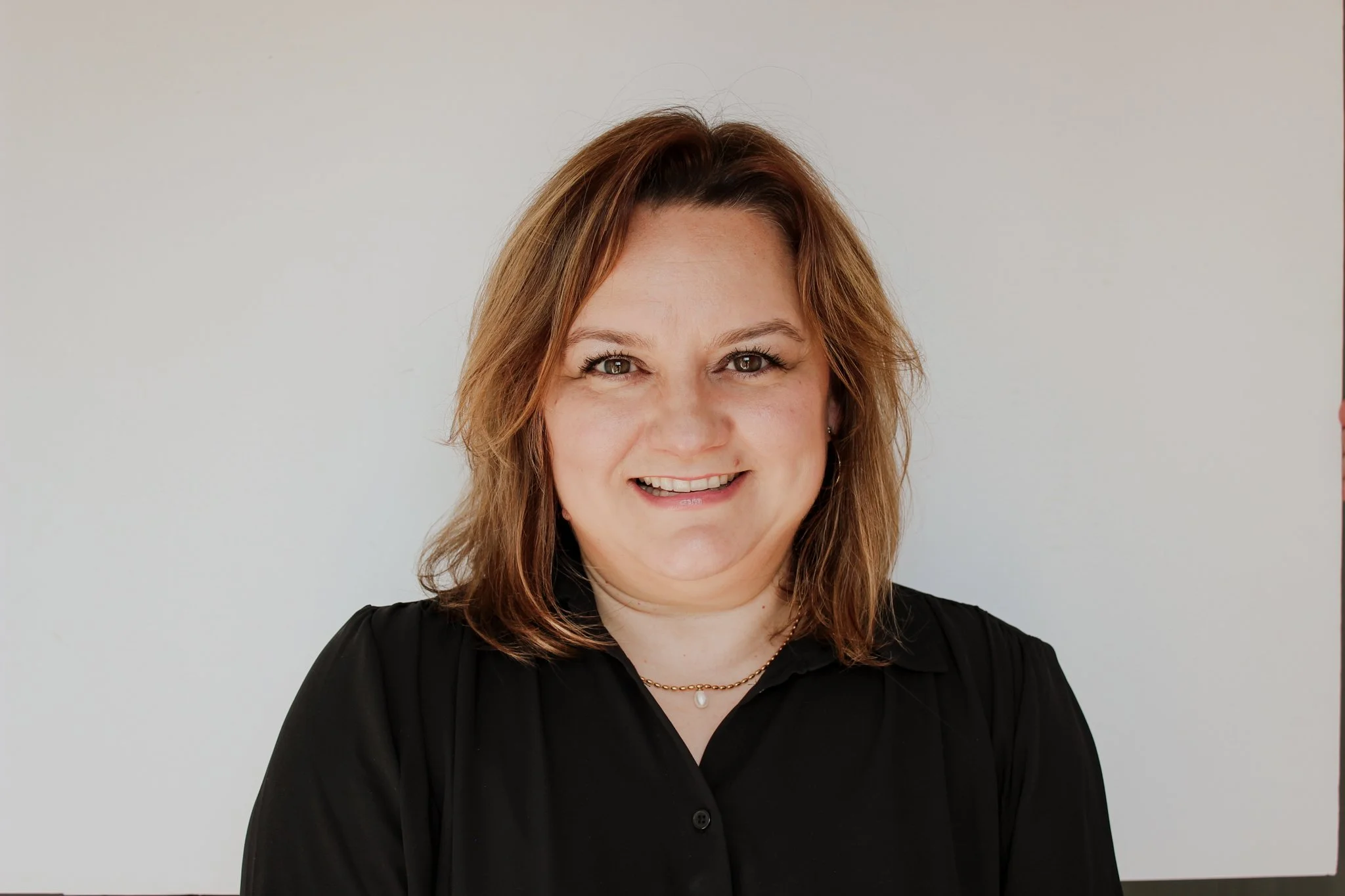 A woman with shoulder-length light brown hair, wearing a black shirt and a pearl necklace, smiling at the camera with a plain light background. Casa del lago's director of children's ministry.