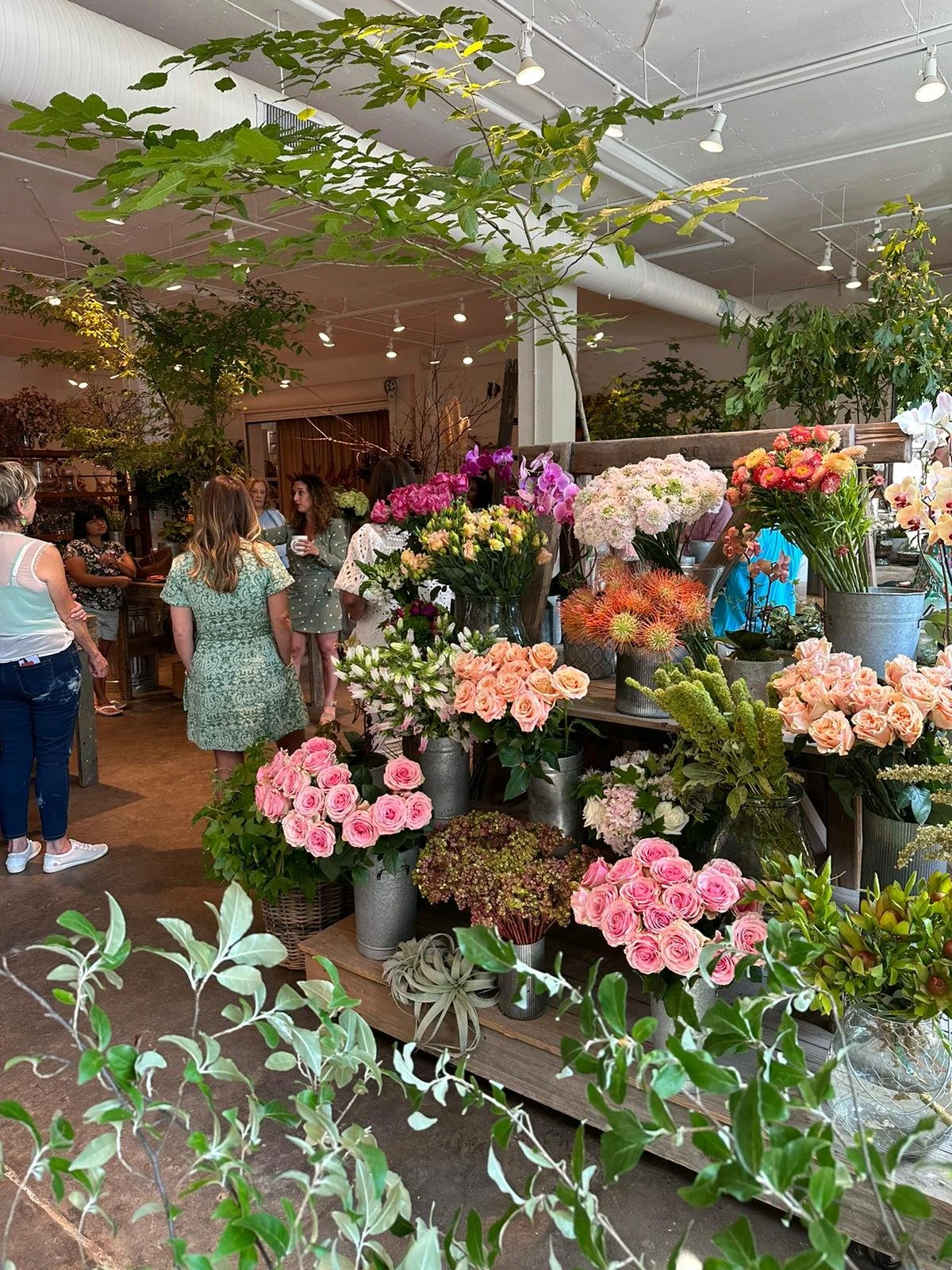 Interior of a flower shop with various colorful flowers on display, including roses and orchids, in vases and on wooden shelves, with people browsing the arrangements.