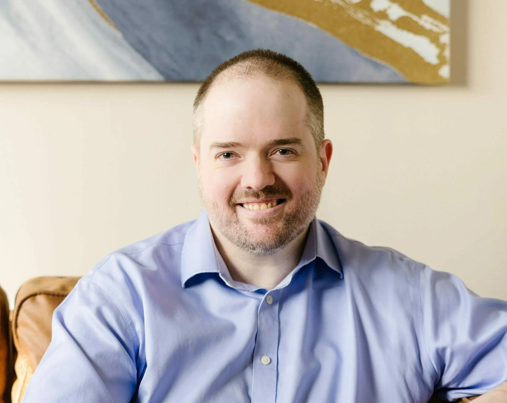A smiling man, David Spielman, with short hair and a beard, wearing a light blue collared shirt, sitting indoors with abstract artwork on the wall behind him.