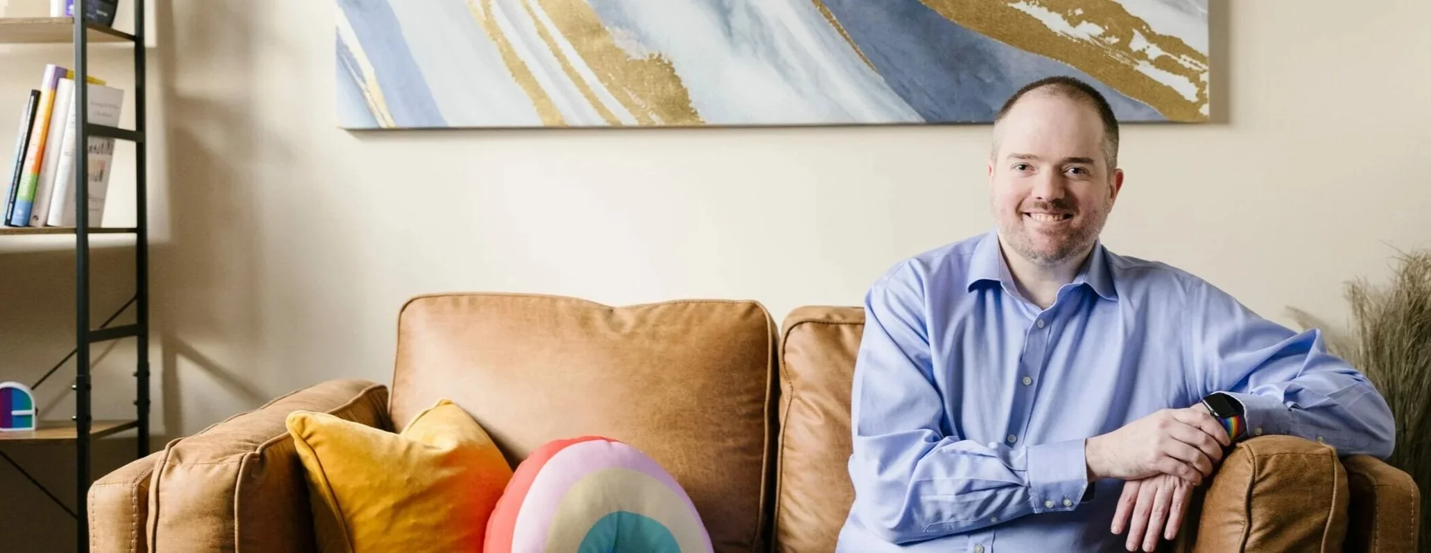 A man, David Spielman, in a blue shirt smiling and sitting on a tan couch with colorful rainbow pillows in a therapy office with a bookshelf and abstract wall art.