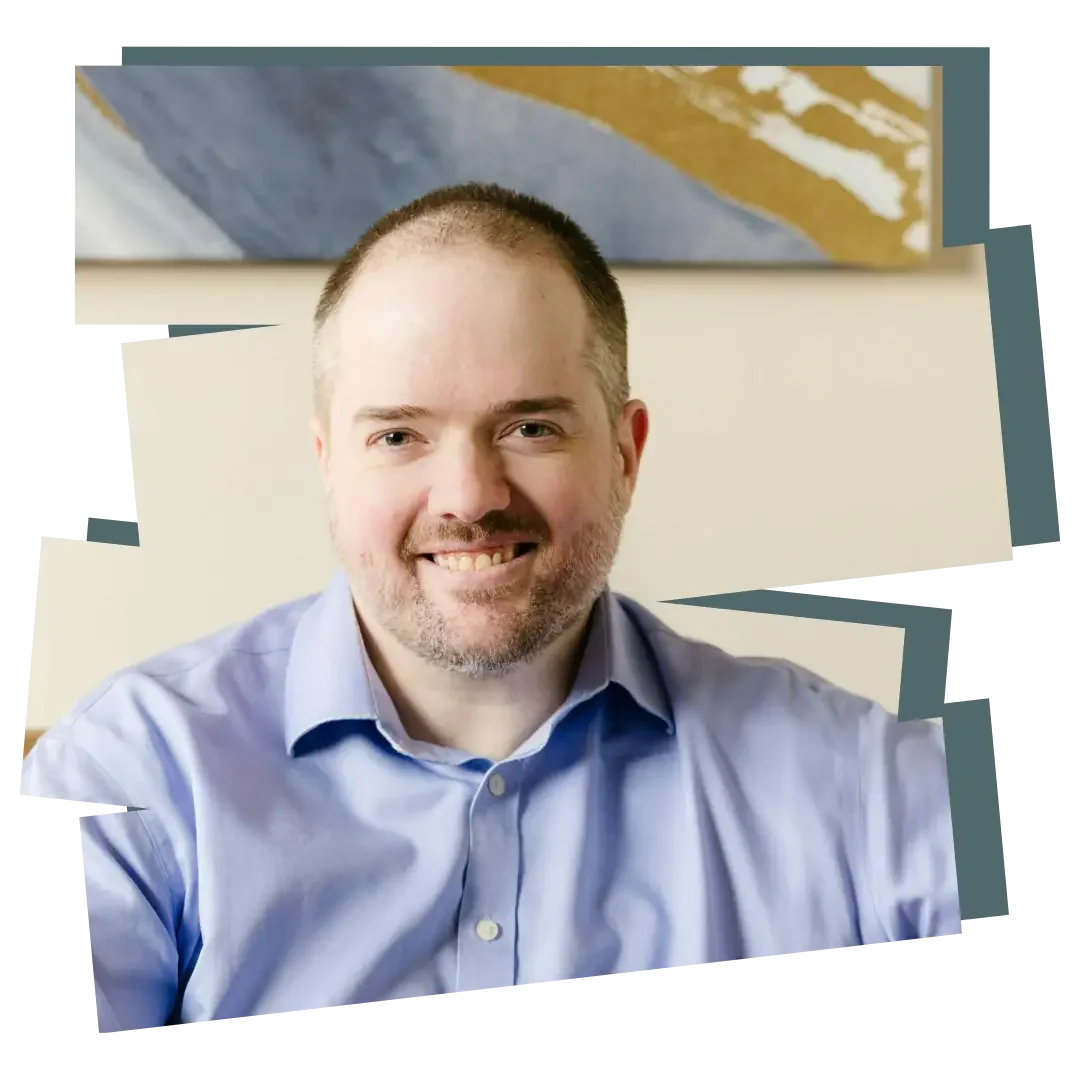 Photo of David Spielman: A smiling man with short hair and a beard, wearing a light blue button-up shirt, sitting indoors with a neutral background and abstract artwork behind him.