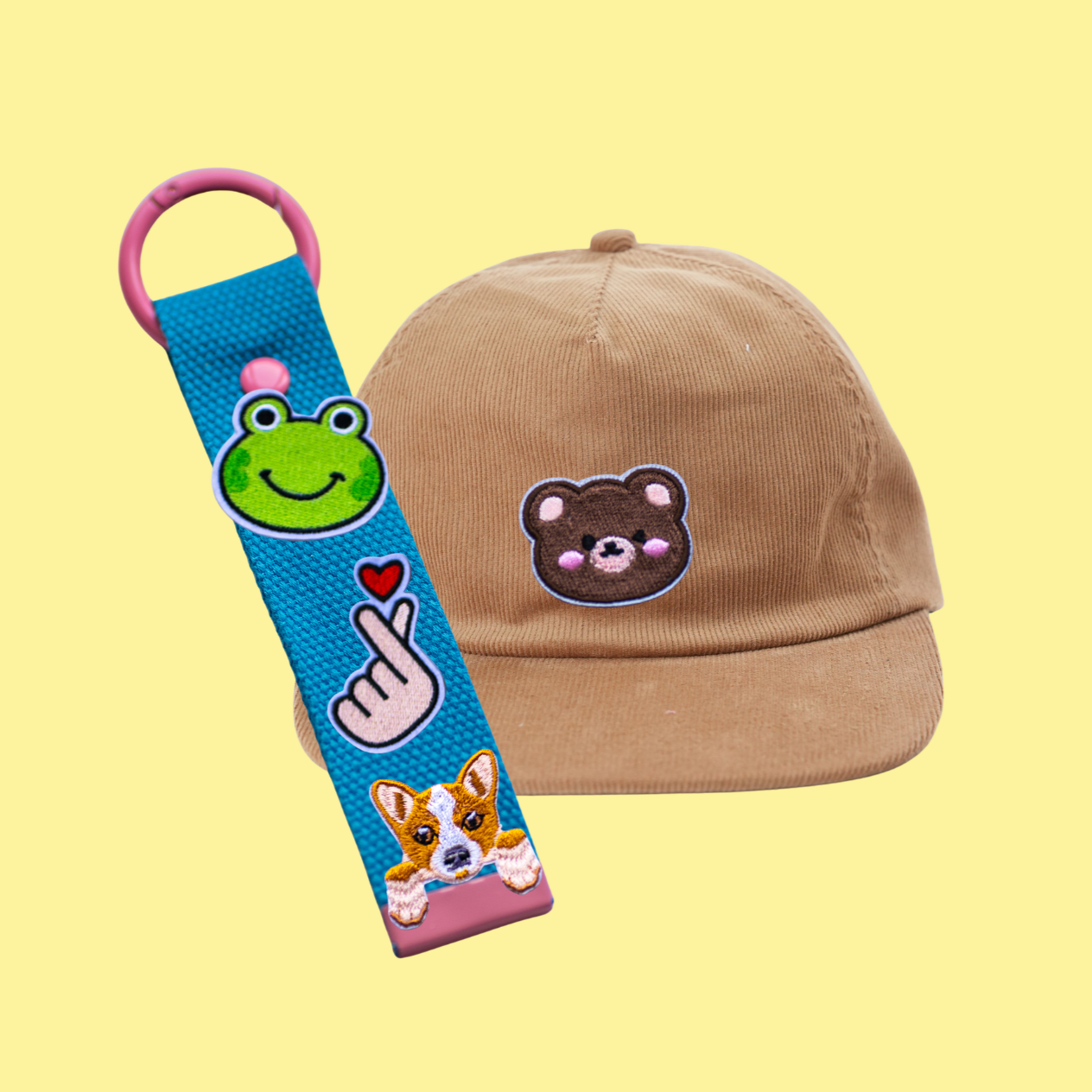 Hellbraune Cap mit einem Stickausschnitt eines Bären, dazu ein Schlüsselanhänger mit verschiedenen bunten Patch-Designs, einschließlich einer grünen Froschwacke, einer Handgeste mit Herz, einem Hundekopf und einem pinken Clip, vor hellem gelbem Hintergrund.