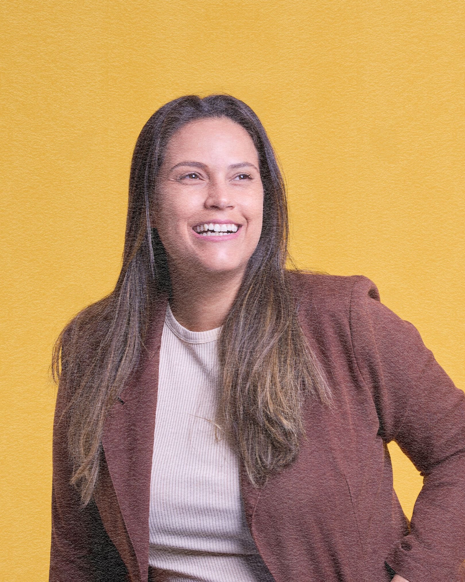 A woman with long brown hair smiling, wearing a brown blazer and a light-colored top, standing against a yellow background.