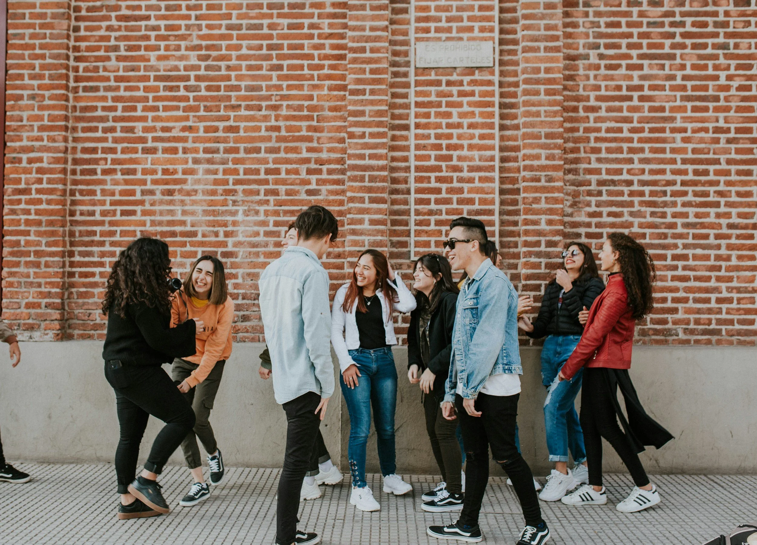 A group of diverse young people laughing and hanging out together outdoors against a brick wall.