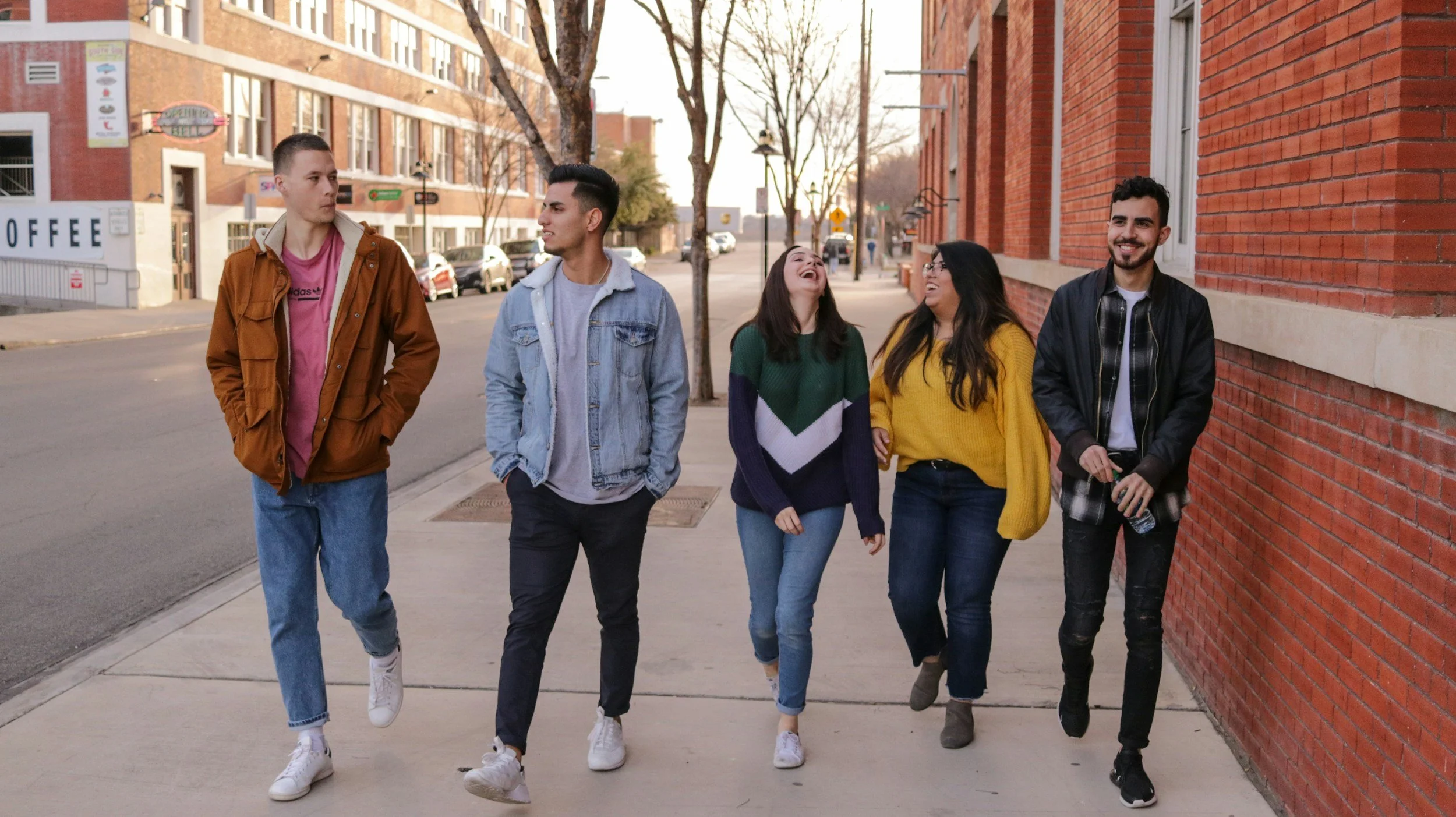 Five diverse young adults walking and talking on a city sidewalk, smiling and laughing, with brick and building background, trees, and parked cars.