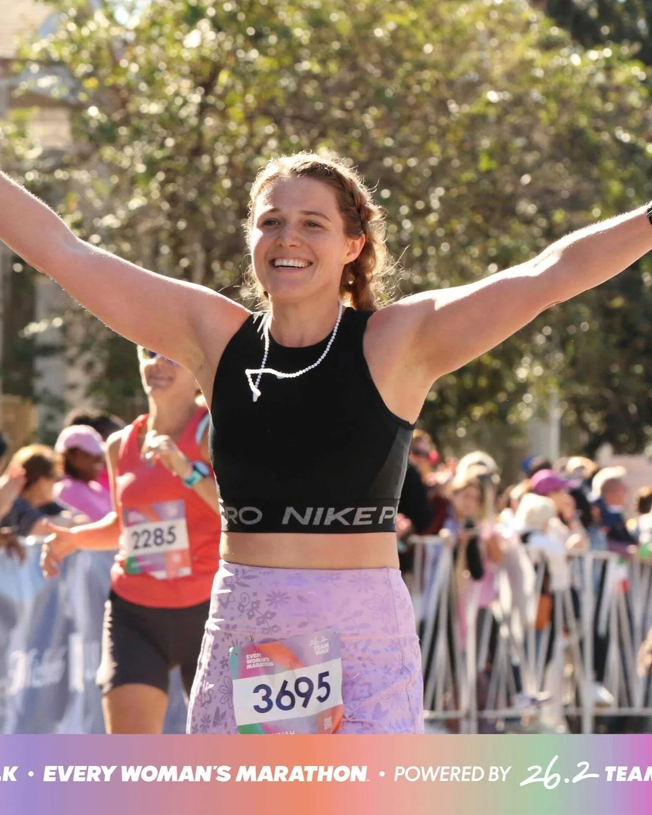 A woman celebrating crossing the finish line at a marathon, wearing a black Ero NikePro sports top, pink patterned shorts, and a race bib numbered 3695, with arms raised and smiling. Other runners and spectators are visible in the background.