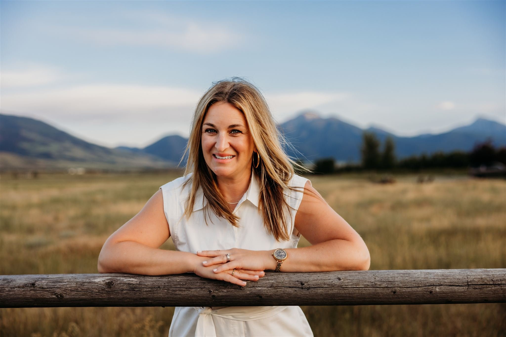 A young woman with shoulder-length brown hair and light eye makeup, smiling at the camera, wearing a cozy black, grey, and red patterned fleece jacket, positioned in a room with red chairs in the background.