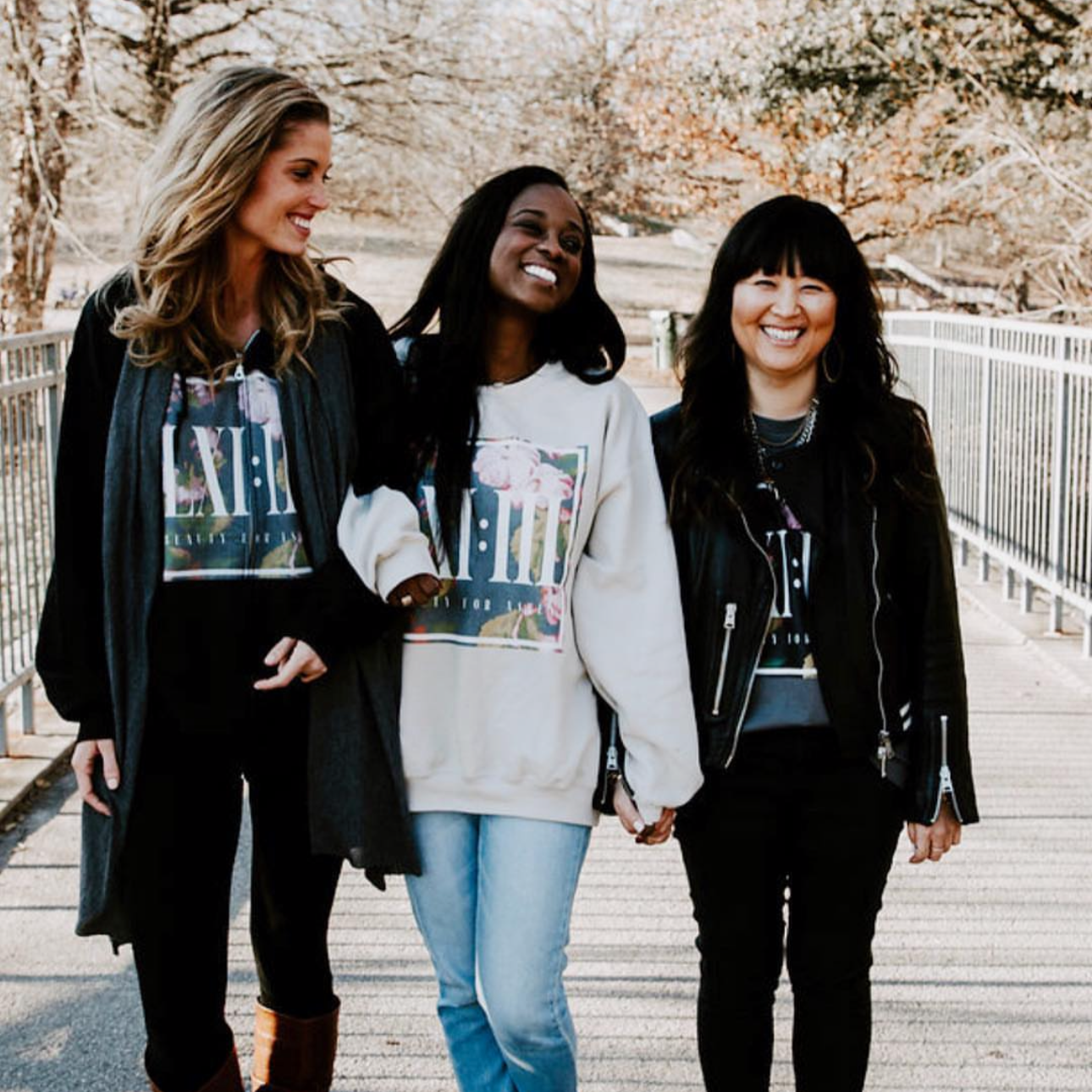 Three women holding hands and smiling on a bridge outdoors in autumn, wearing casual jackets.