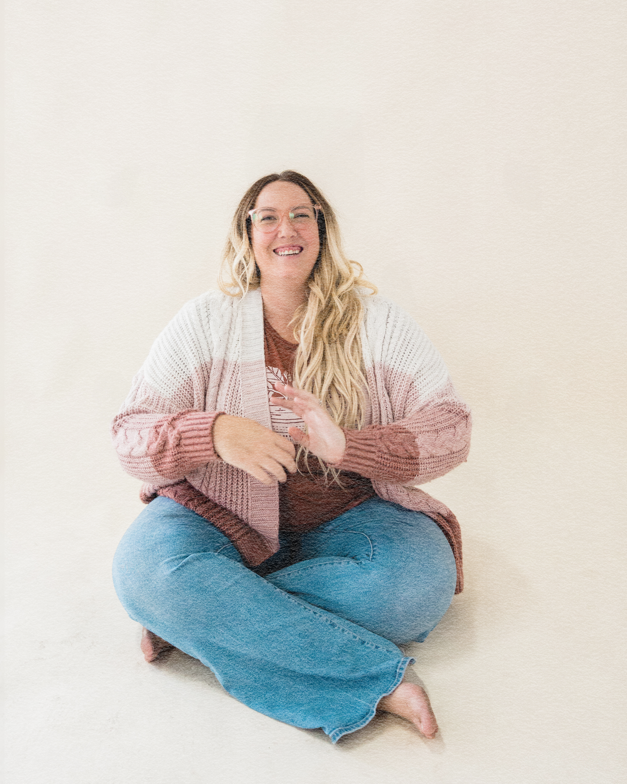 A woman with long blonde hair, glasses, and a wide smile, sitting cross-legged on the floor against a plain light-colored background, wearing a patterned sweater and blue jeans.