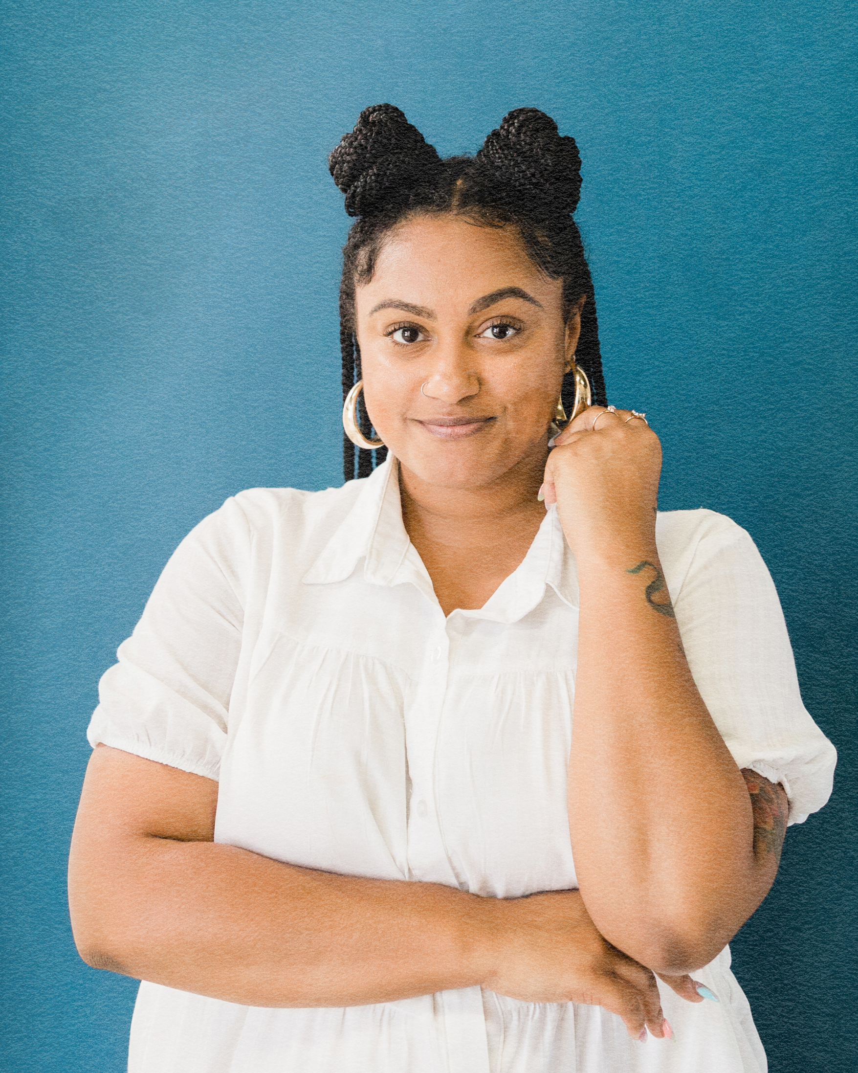 A woman with braided hair styled in space buns, wearing hoop earrings and a white button-up shirt, stands against a blue background with her arm across her chest and a confident expression.