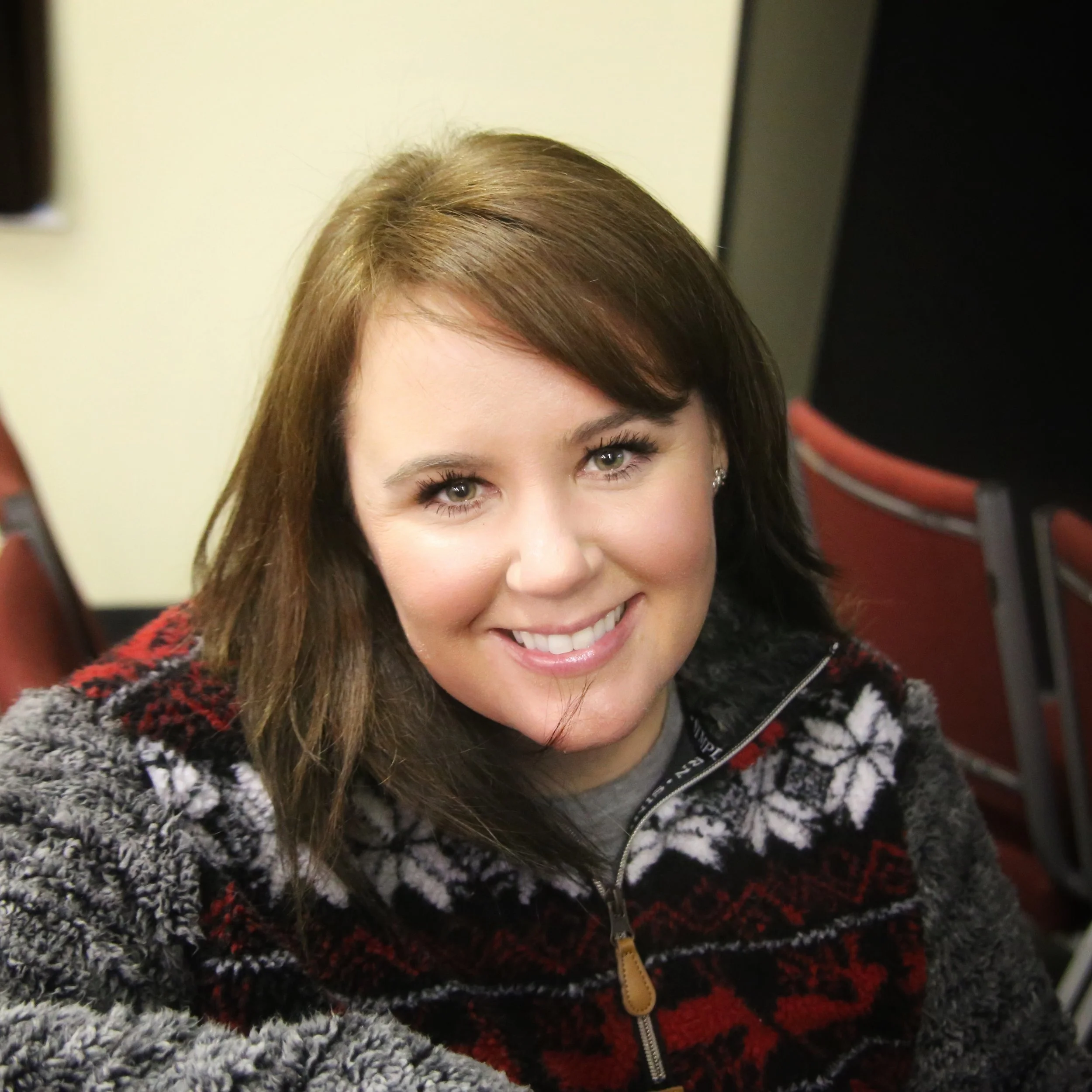 A young woman with shoulder-length brown hair and light eye makeup, smiling at the camera, wearing a cozy black, grey, and red patterned fleece jacket, positioned in a room with red chairs in the background.