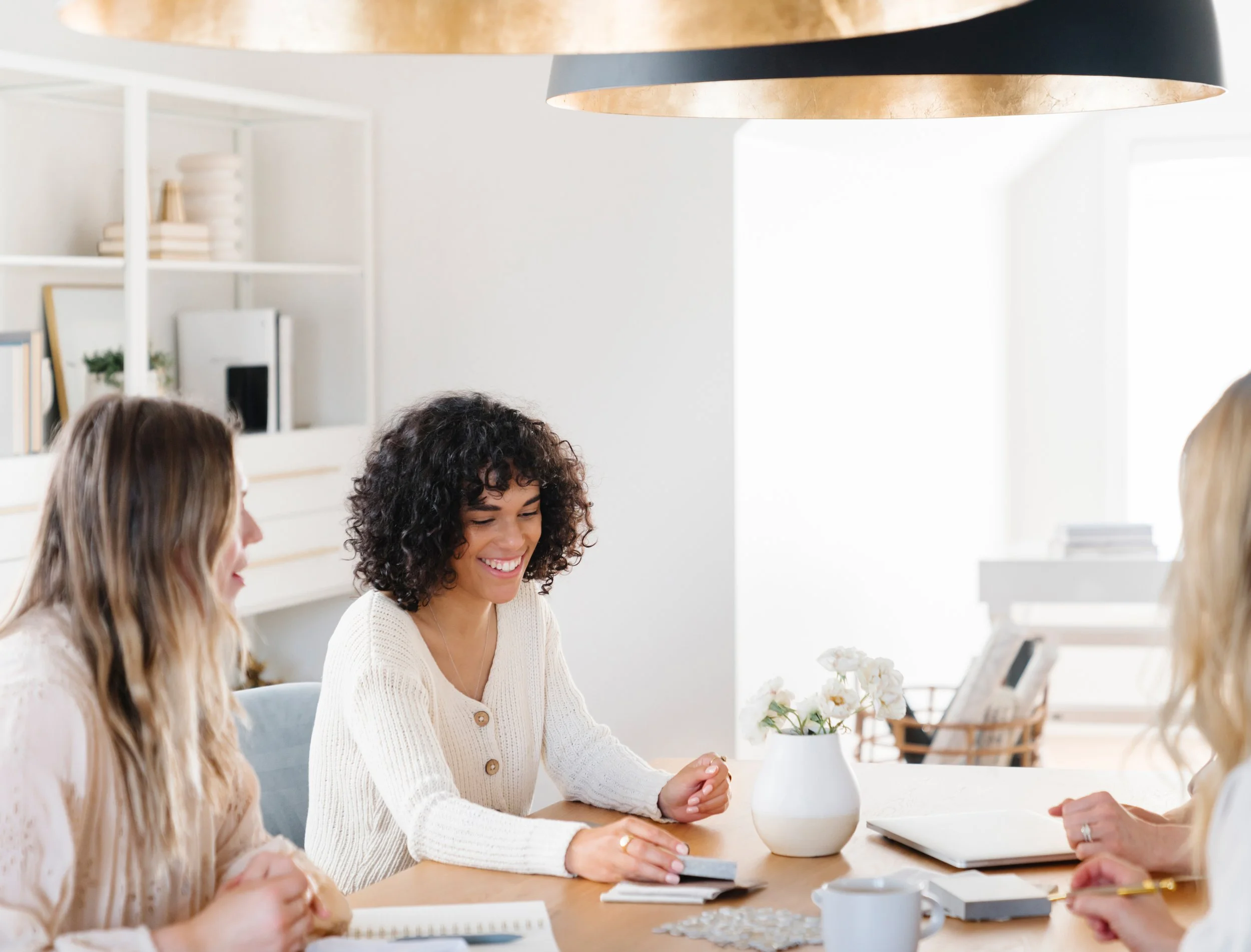 Four women sitting at a table in a bright, modern office or meeting room, engaged in conversation and smiling, with notebooks, a laptop, and a coffee mug on the table.