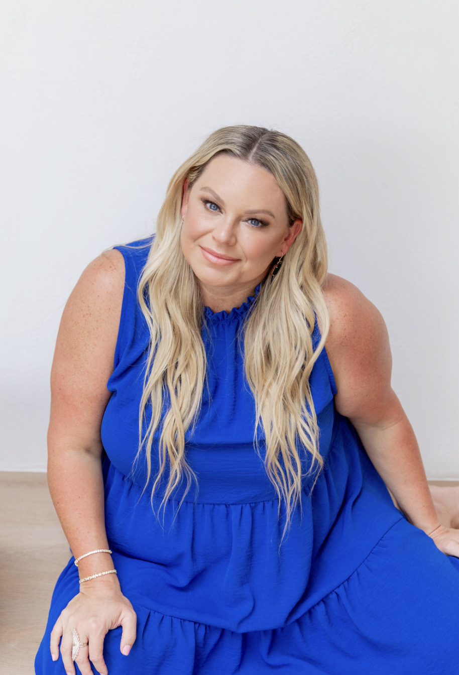A woman with long blonde hair wearing a sleeveless royal blue dress, sitting against a white wall, looking at the camera with a slight smile.