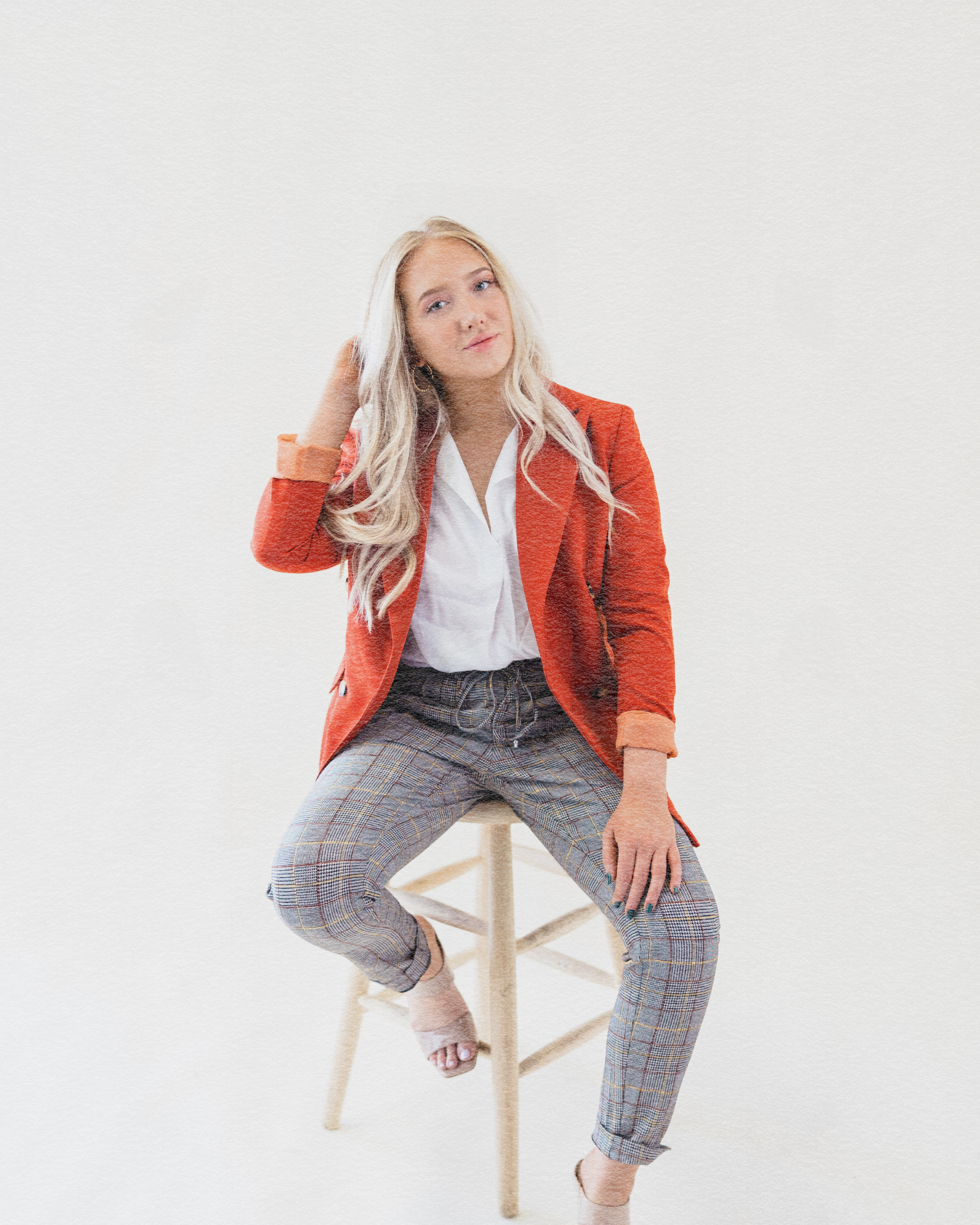 A young woman with long blond hair sitting on a light wooden stool against a plain white background, wearing a red blazer, white shirt, and plaid pants, with relaxed pose and thoughtful expression.
