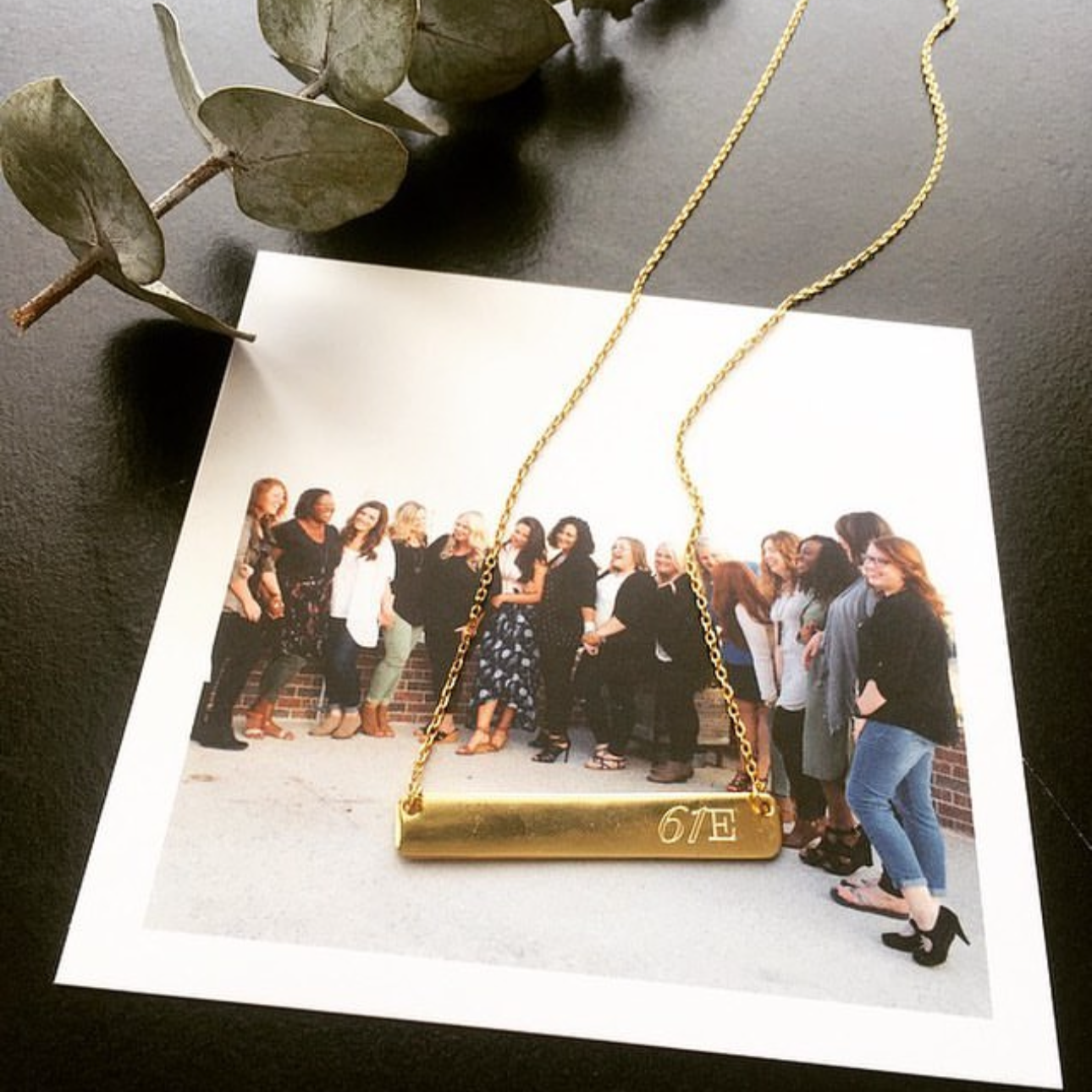 A group photo of women standing in a line, dressed in casual and semi-formal attire, taken outdoors against a brick wall. There is a gold necklace with a bar pendant marked '2019' placed on top of the photo, with a small branch of eucalyptus leaves nearby.