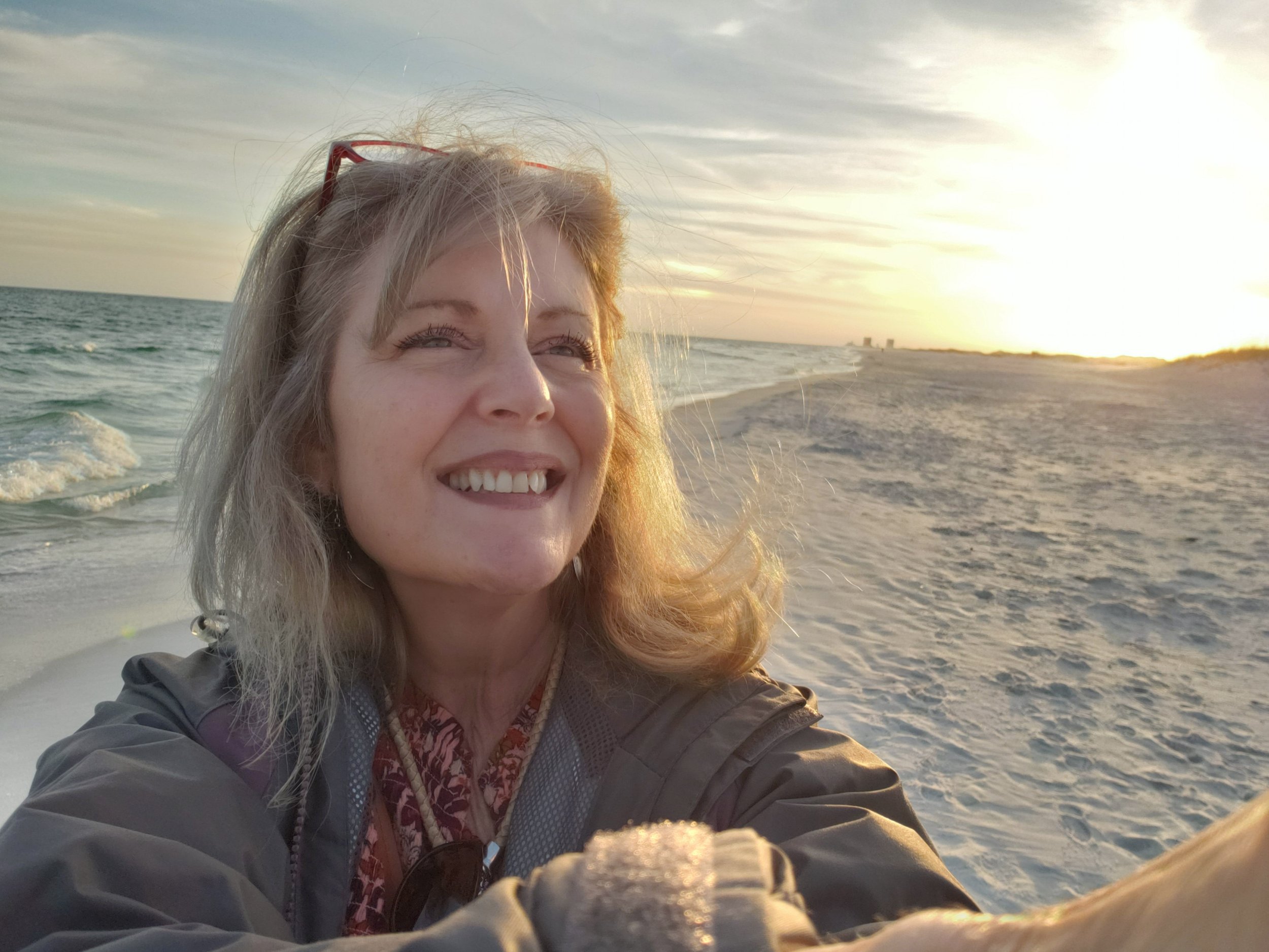 A woman with blonde hair smiling as she takes a selfie on a sandy beach during sunset, with the ocean waves behind her.