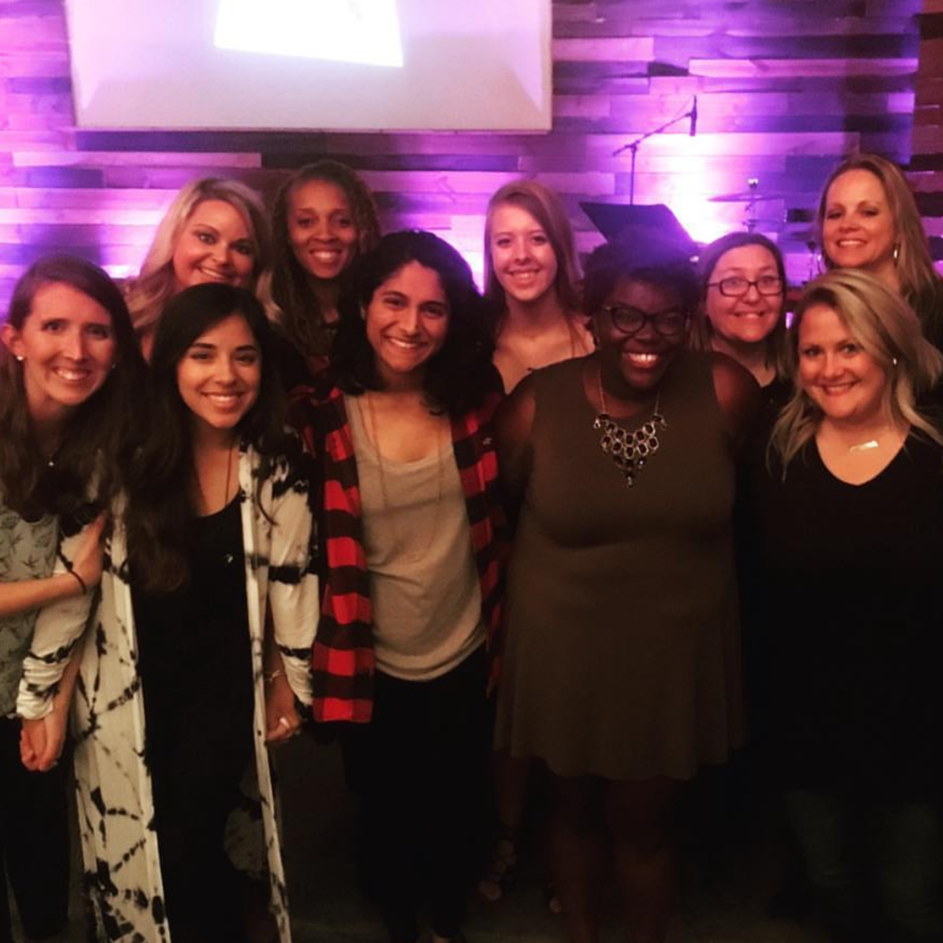 Group of women smiling at an indoor event with purple lighting and a wooden wall background, one woman wearing a graduation cap.