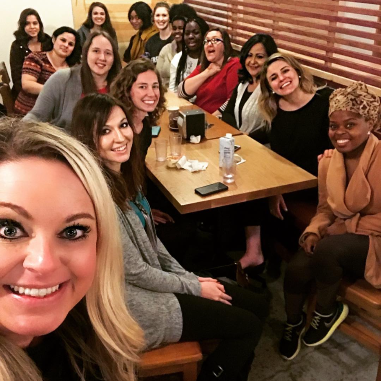 A group of women gathered around a large wooden table in a restaurant, smiling for a selfie, with some drinking water and snacks on the table.