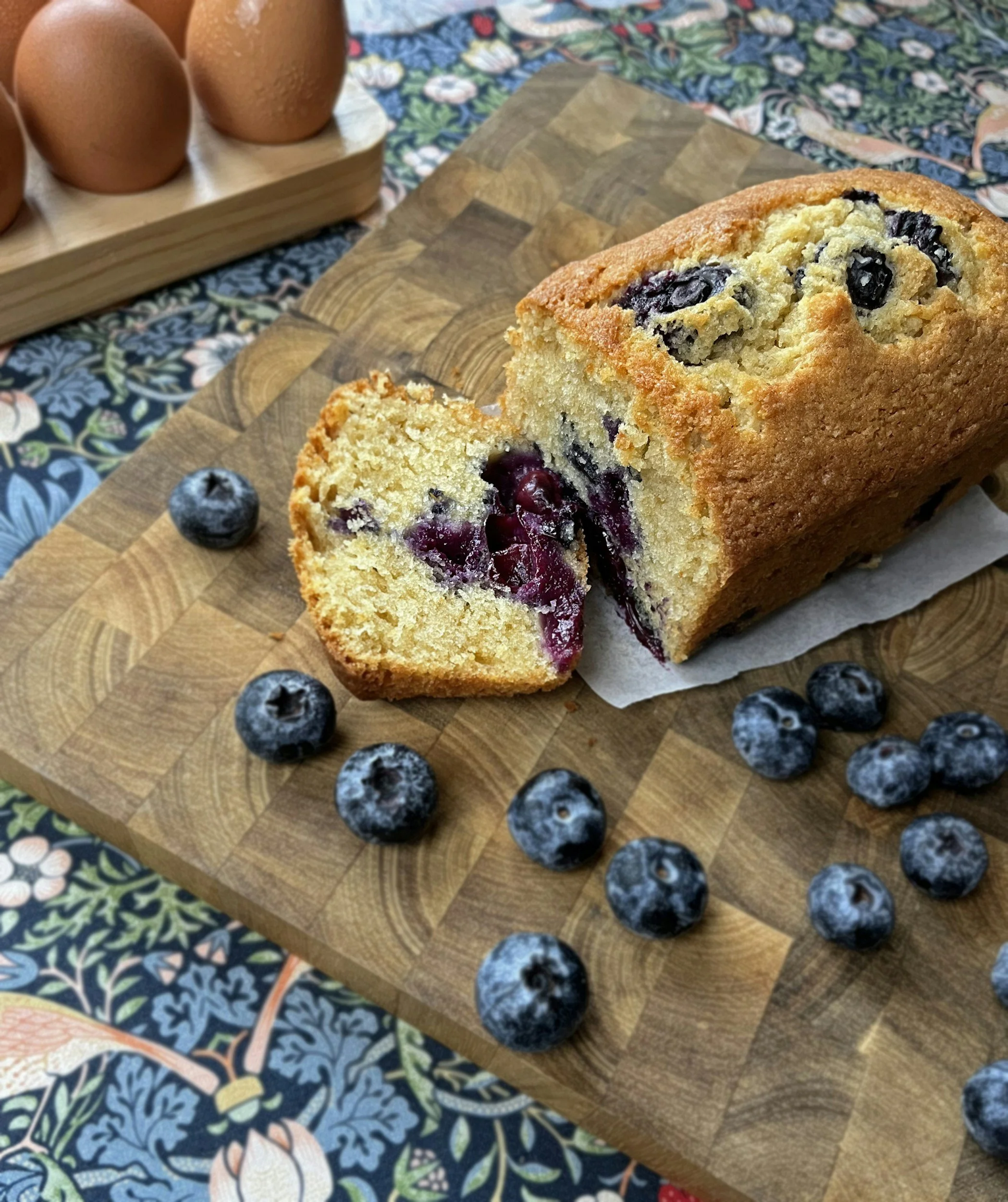 A freshly baked blueberry loaf cake on a wooden cutting board, surrounded by scattered blueberries, with a dozen brown eggs in a wooden holder in the background, all placed on a floral patterned tablecloth.