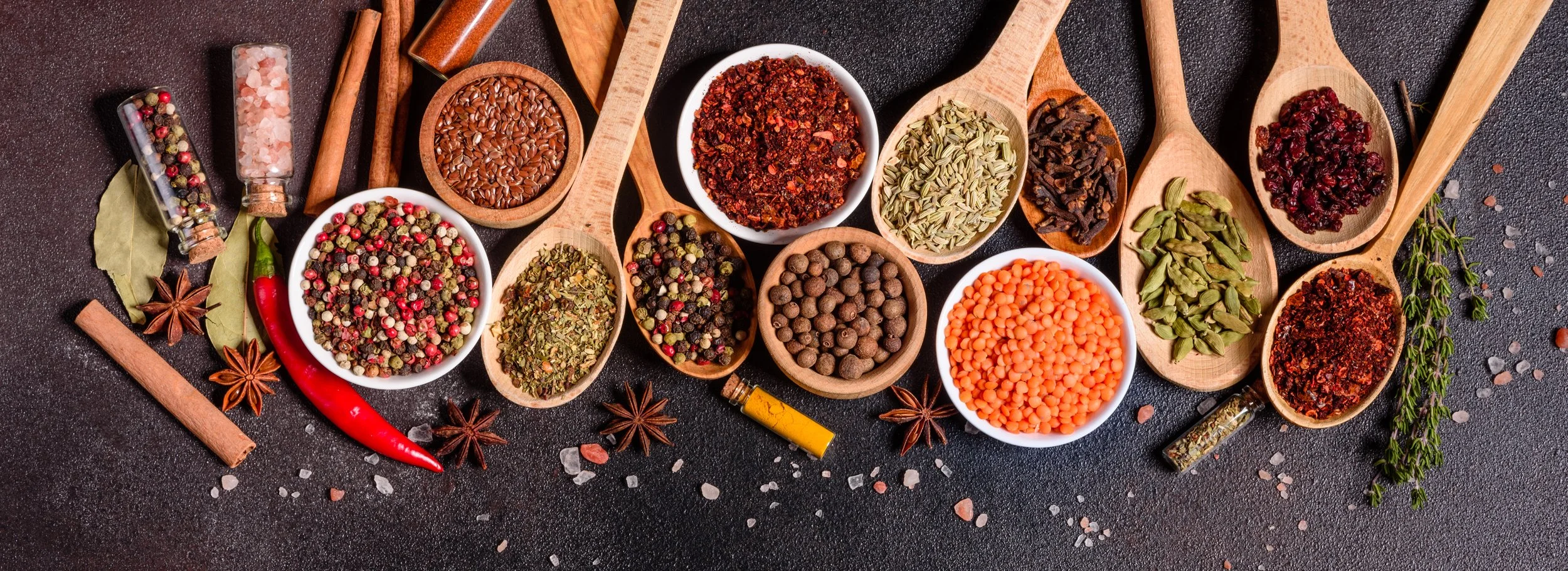 An assortment of colorful spices and herbs in small bowls and wooden spoons on a black surface, including red pepper flakes, black pepper, cayenne, cinnamon sticks, bay leaves, star anise, paprika, and dried herbs.