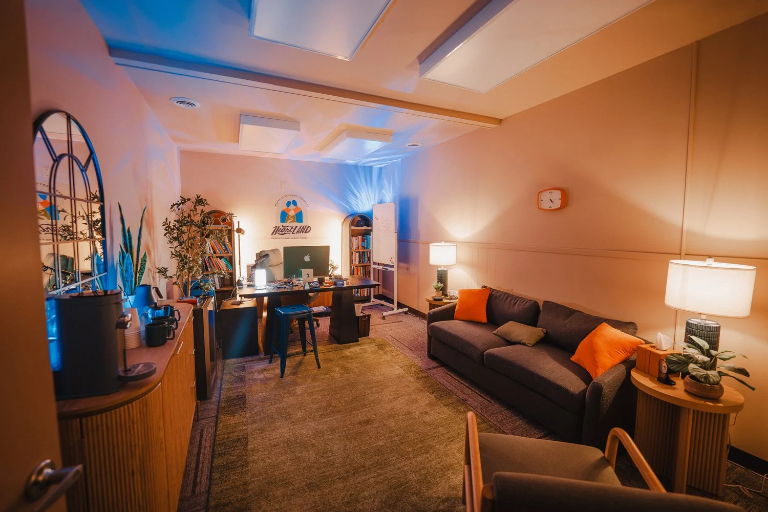 A cozy office or lounge room with warm lighting, a dark sofa with orange and green pillows, a side table with lamp and plants, a large wooden desk with a computer, and bookshelves. There are plants, lamps, and a whiteboard, with a logo reading "HeartLAND" on the wall.