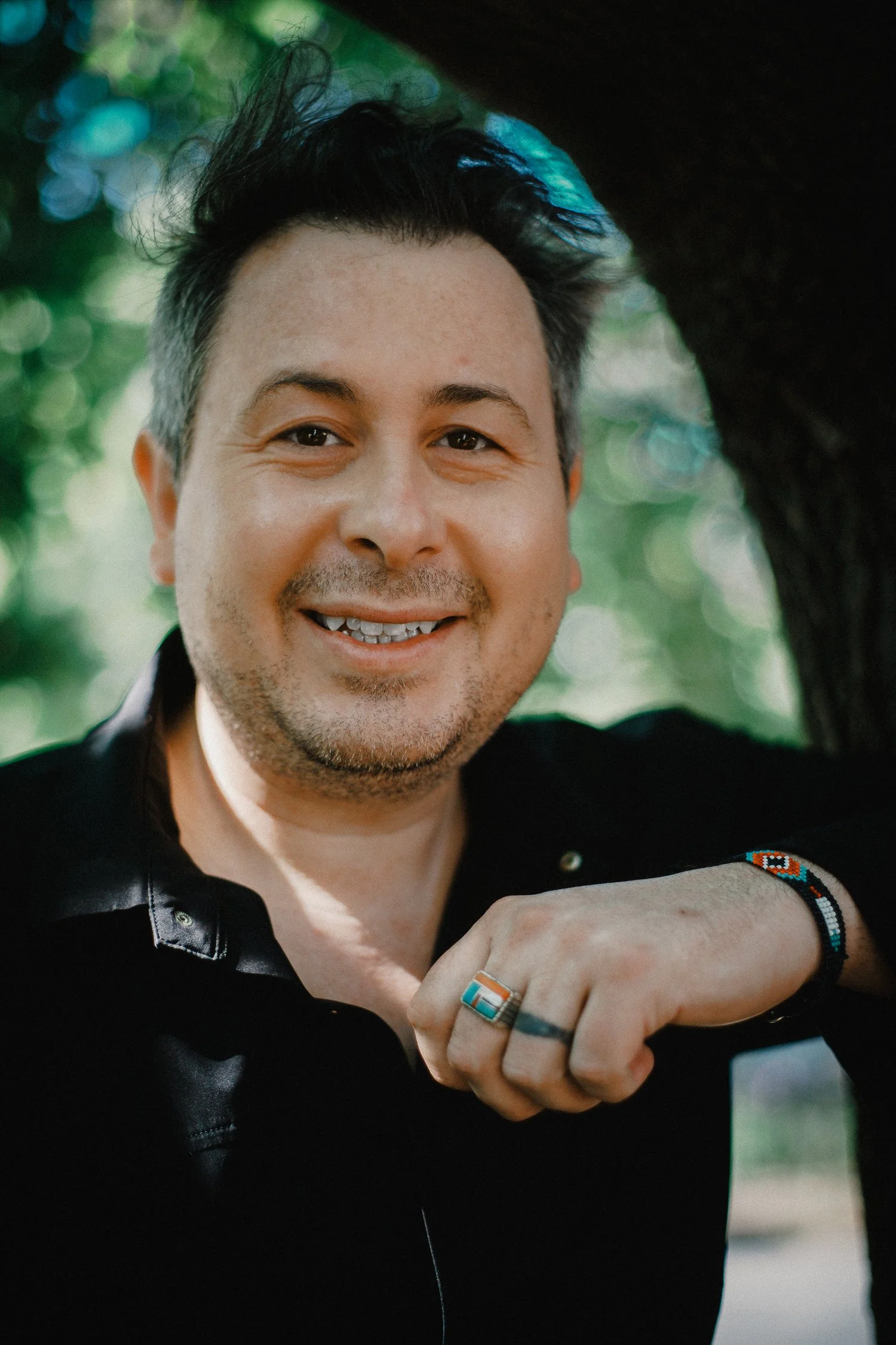 Close-up of smiling man with short dark hair, wearing rings and beaded bracelets, dressed in black leather jacket, outdoors with trees in background.