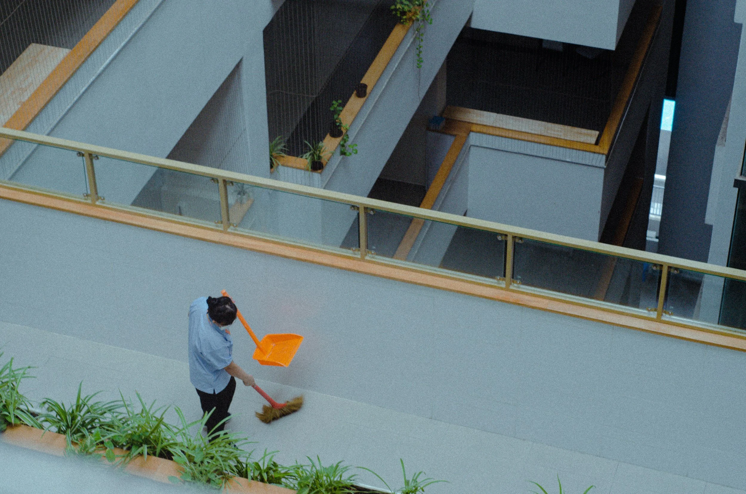 A person sweeping with a broom in a modern building with multiple floors, glass railings, and potted plants.