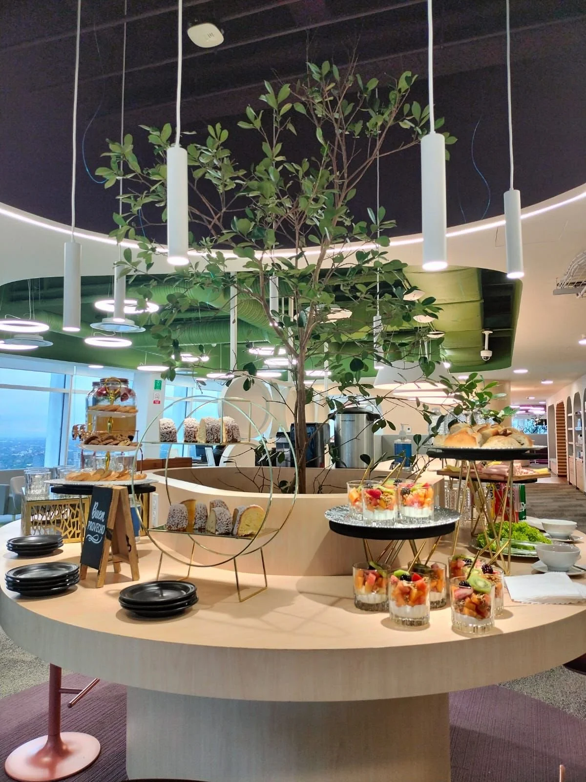 Mesa con diversos postres y snacks en un ambiente interior con un árbol decorativo en el centro, iluminado por luces modernas, en medio de un espacio que parece ser un restaurante o cafetería.