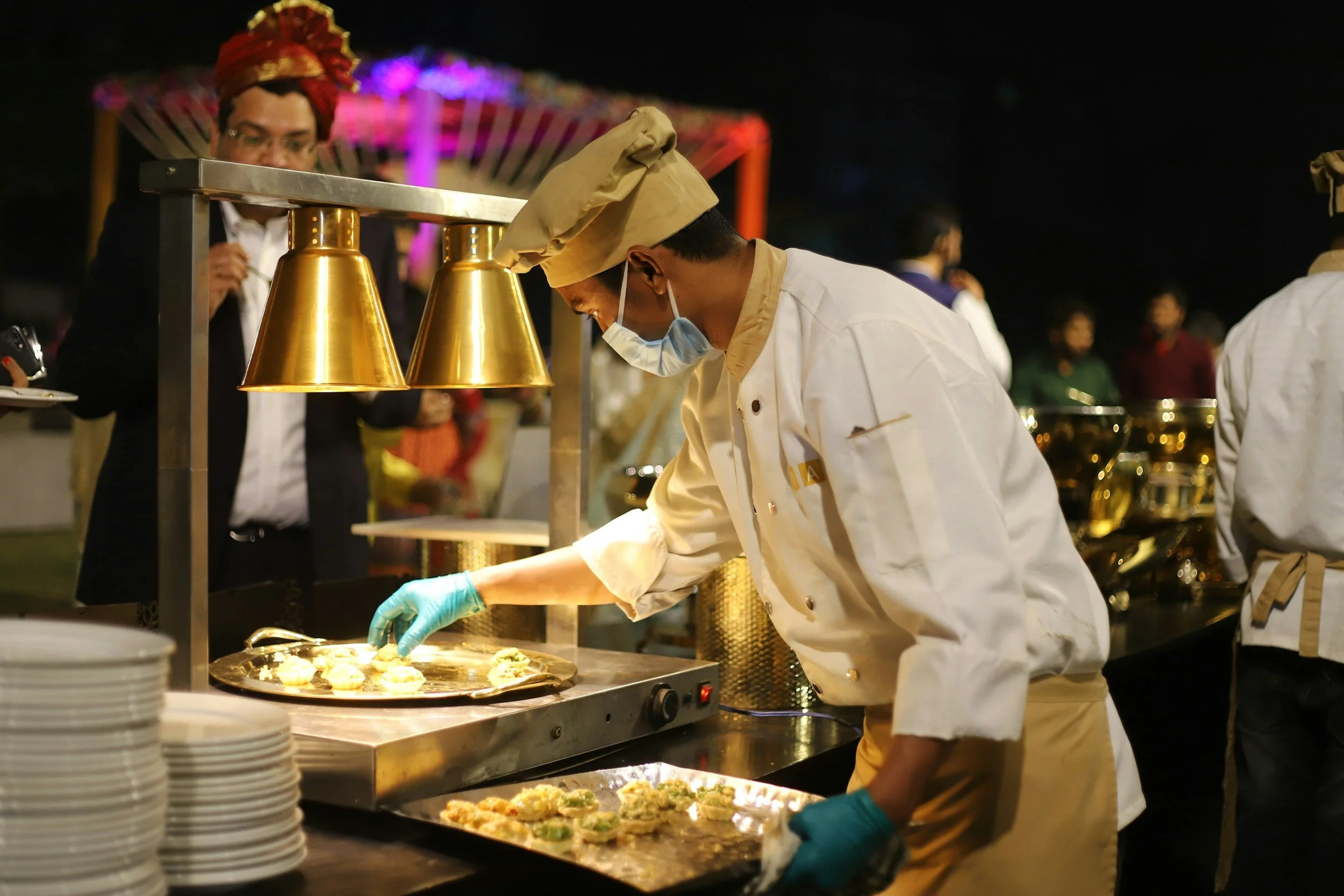 Un chef preparando comida en una feria nocturna con luces de colores y otras personas en el fondo.