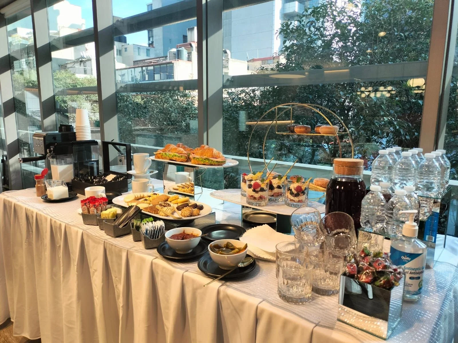 Mesa con desayuno tipo buffet con croissants, sandwiches, fruta, dulces, té, agua y productos de higiene, en un espacio con ventanas grandes y vista a edificios y árboles.